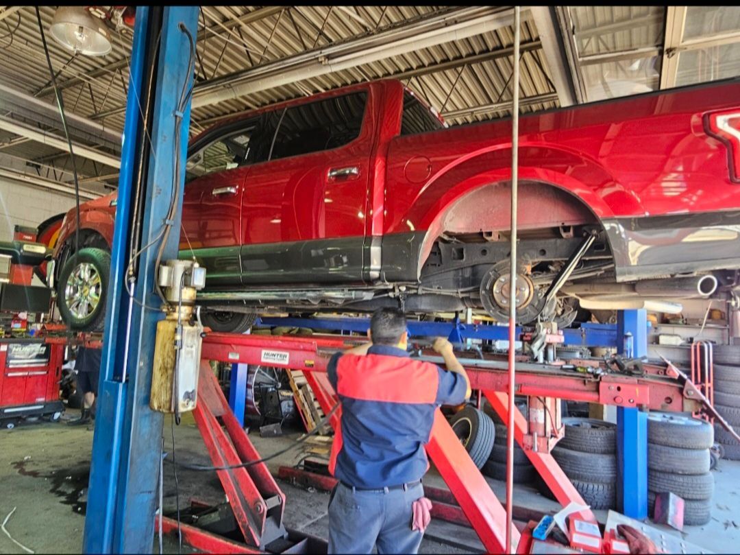 A technician works on the undercarriage of a red pickup truck elevated on a hydraulic lift inside an auto repair shop.