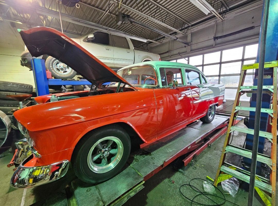 A bright red vintage car with its hood open, parked on a service lift inside an automotive repair shop.