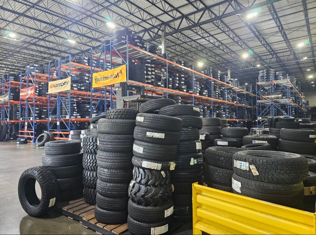Stacked tires in a large warehouse with rows of metal shelving filled with tire inventory.