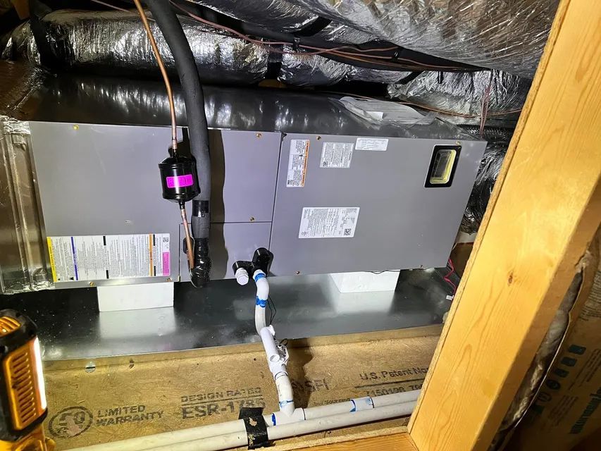An HVAC air handler unit installed in an attic space, with connected insulated ductwork and a white PVC drain pipe.