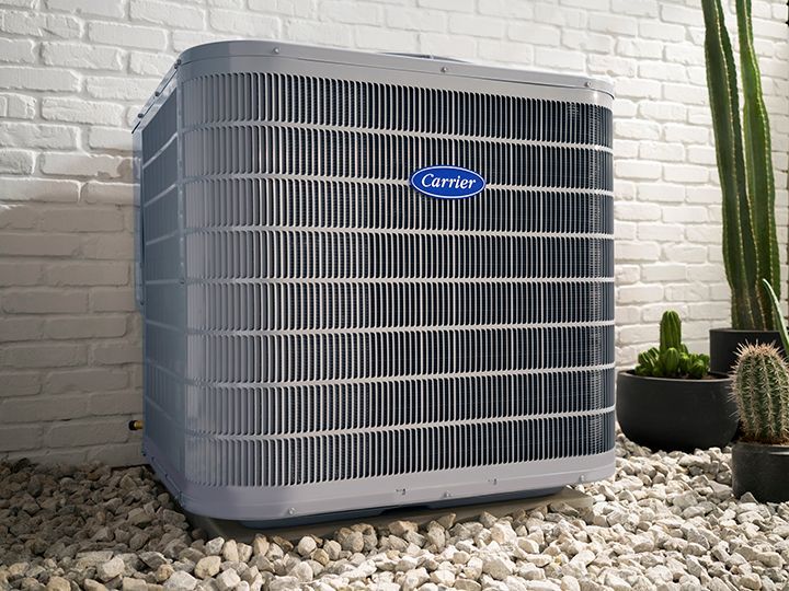 A grey Carrier air conditioning unit sits outdoors on a bed of white gravel against a white brick wall with potted cacti.