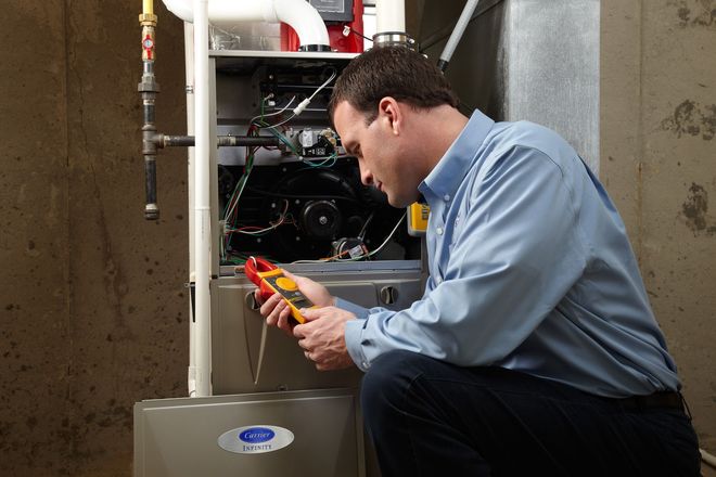 A technician kneels in a basement, using a digital multimeter to test the electrical components of a furnace.