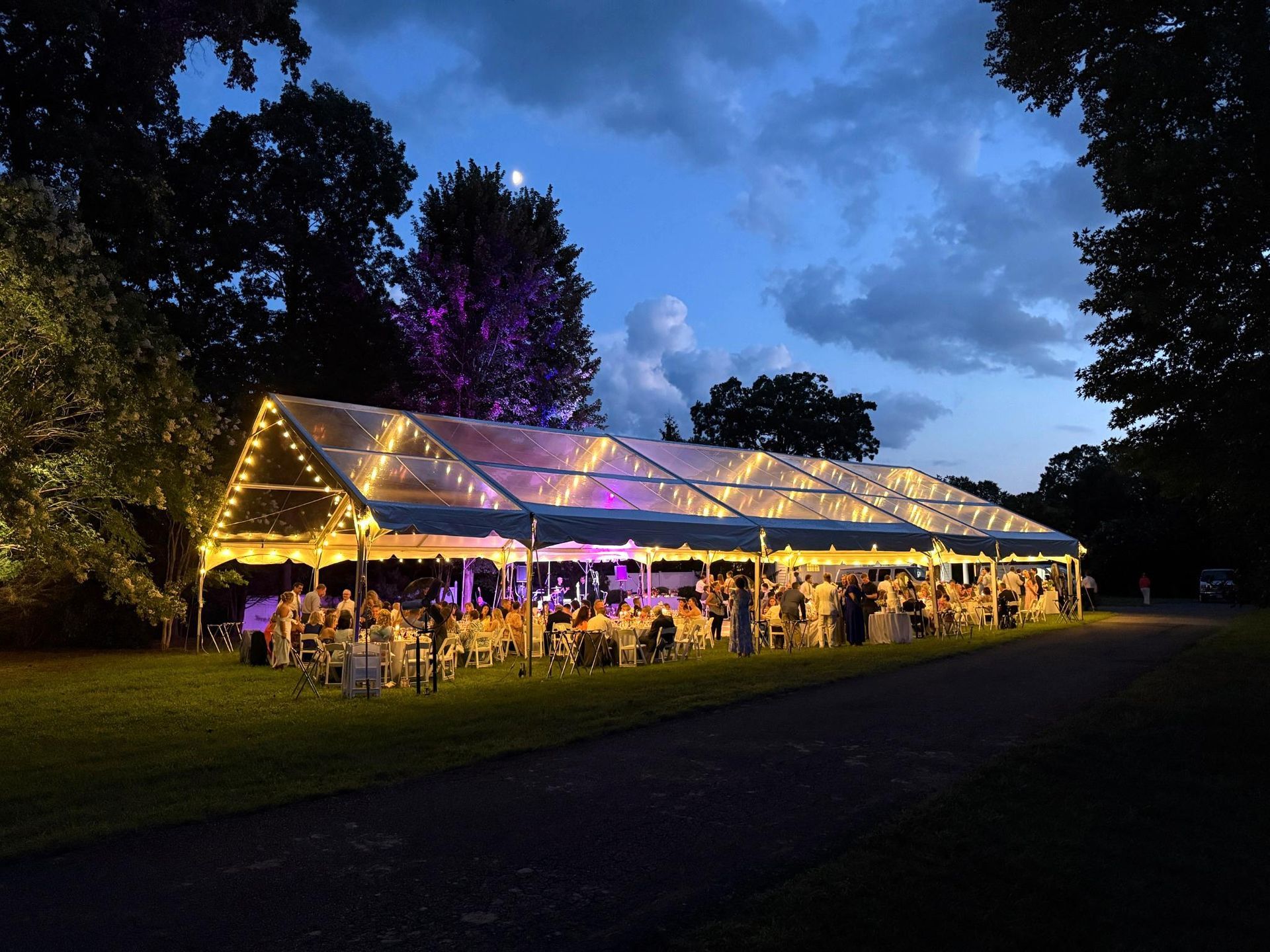 Lit-up tent at night, hosting a party on a grassy lawn with guests, string lights, and trees in the background.