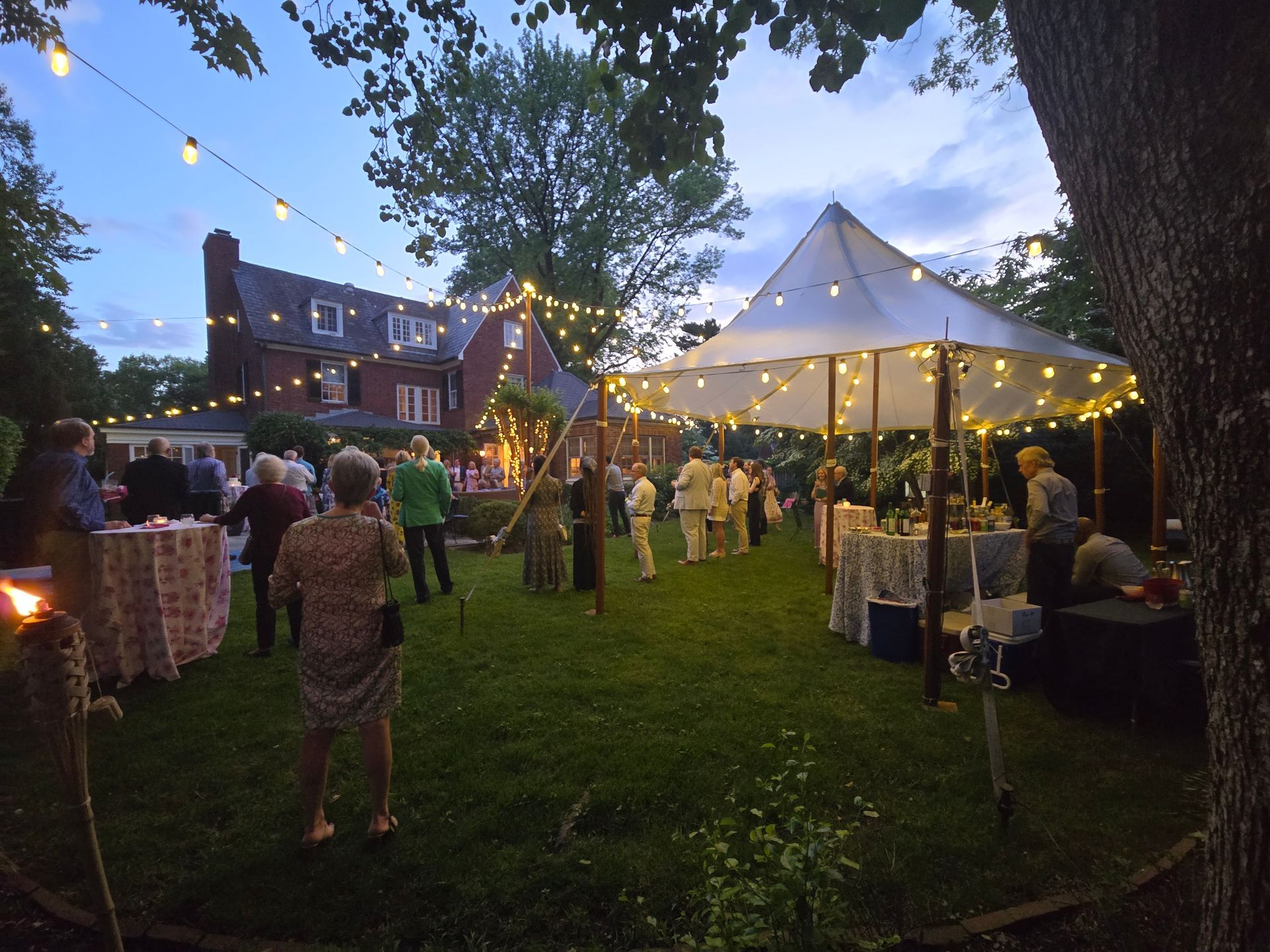 Evening outdoor party with guests, tables, strung lights, and a tent set up on a lawn.