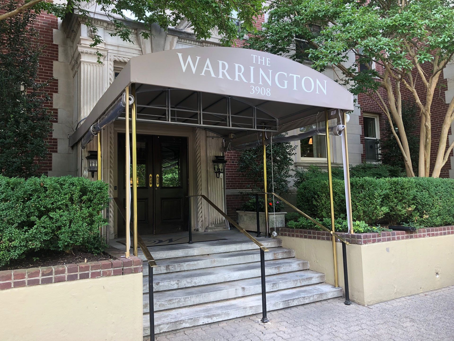 The Warrington apartment building entrance with steps, a brown awning, and greenery.