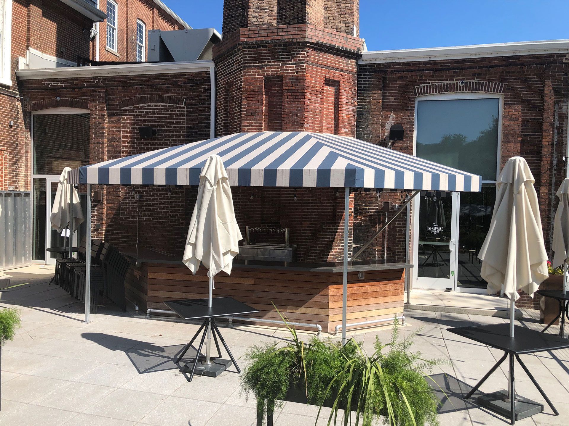 Outdoor patio with striped canopy, brick building backdrop, tables, and plants.