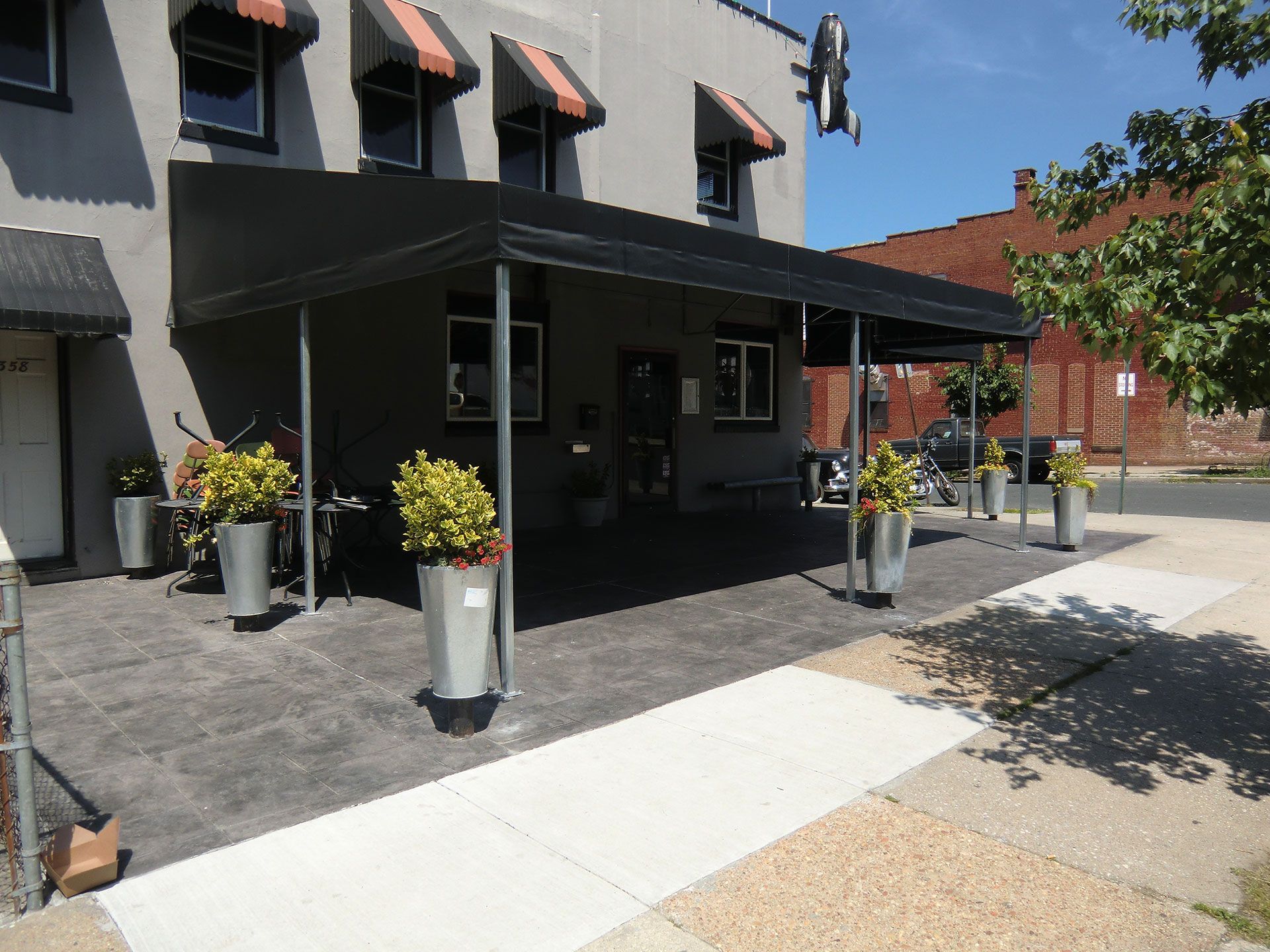 Gray building with black awning, outdoor seating, potted plants. Sunny day.