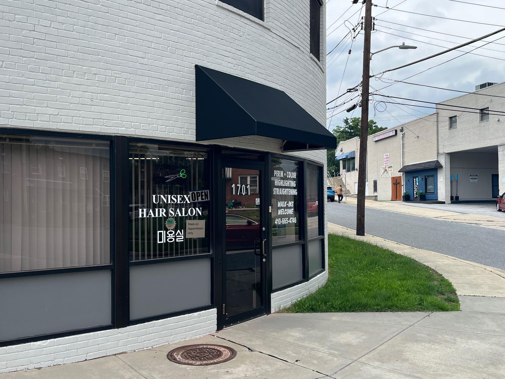 A brick building with a black awning houses a unisex hair salon; glass windows, front door, and a grassy area.
