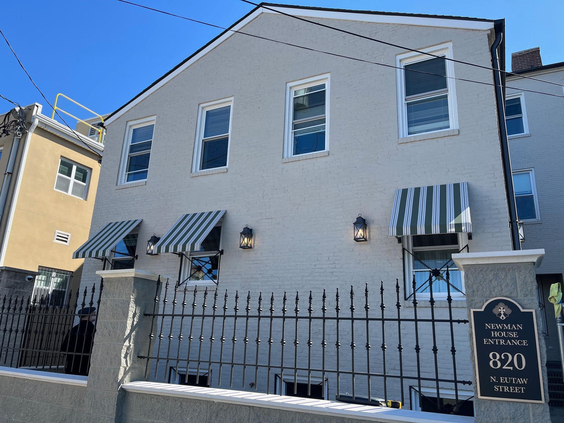 Two-story grey building with awnings over windows. Wrought-iron fence. Sign reads 