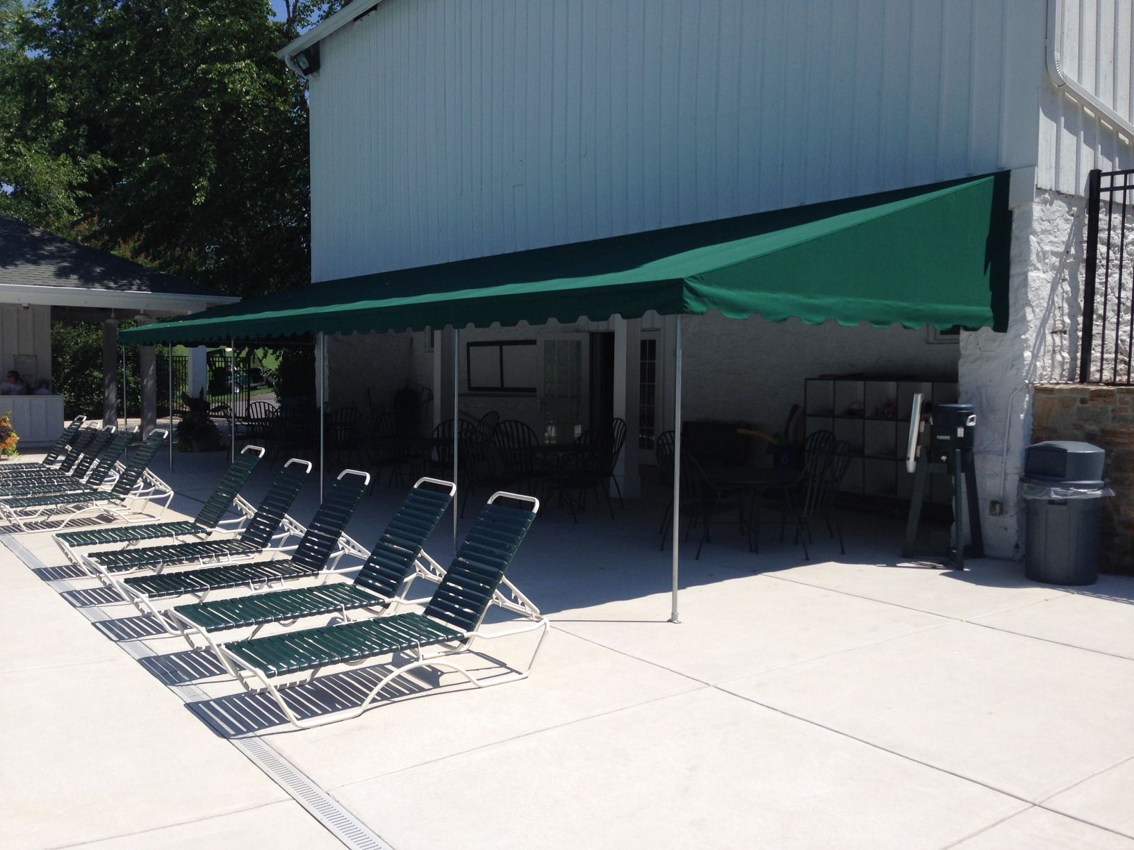 Green awning over pool chairs near a white building.