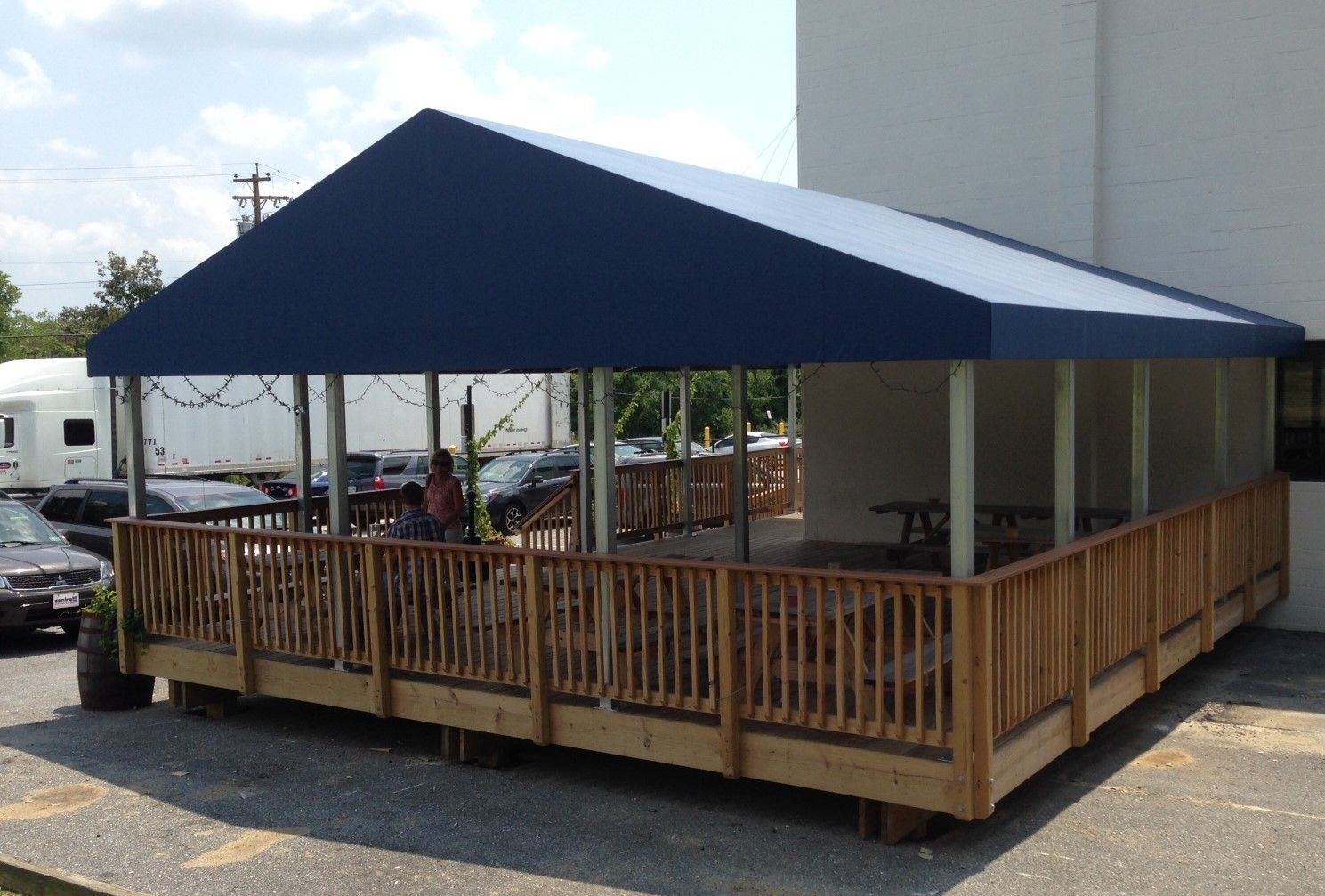Outdoor wooden patio with blue awning; tables and chairs, next to a white building.