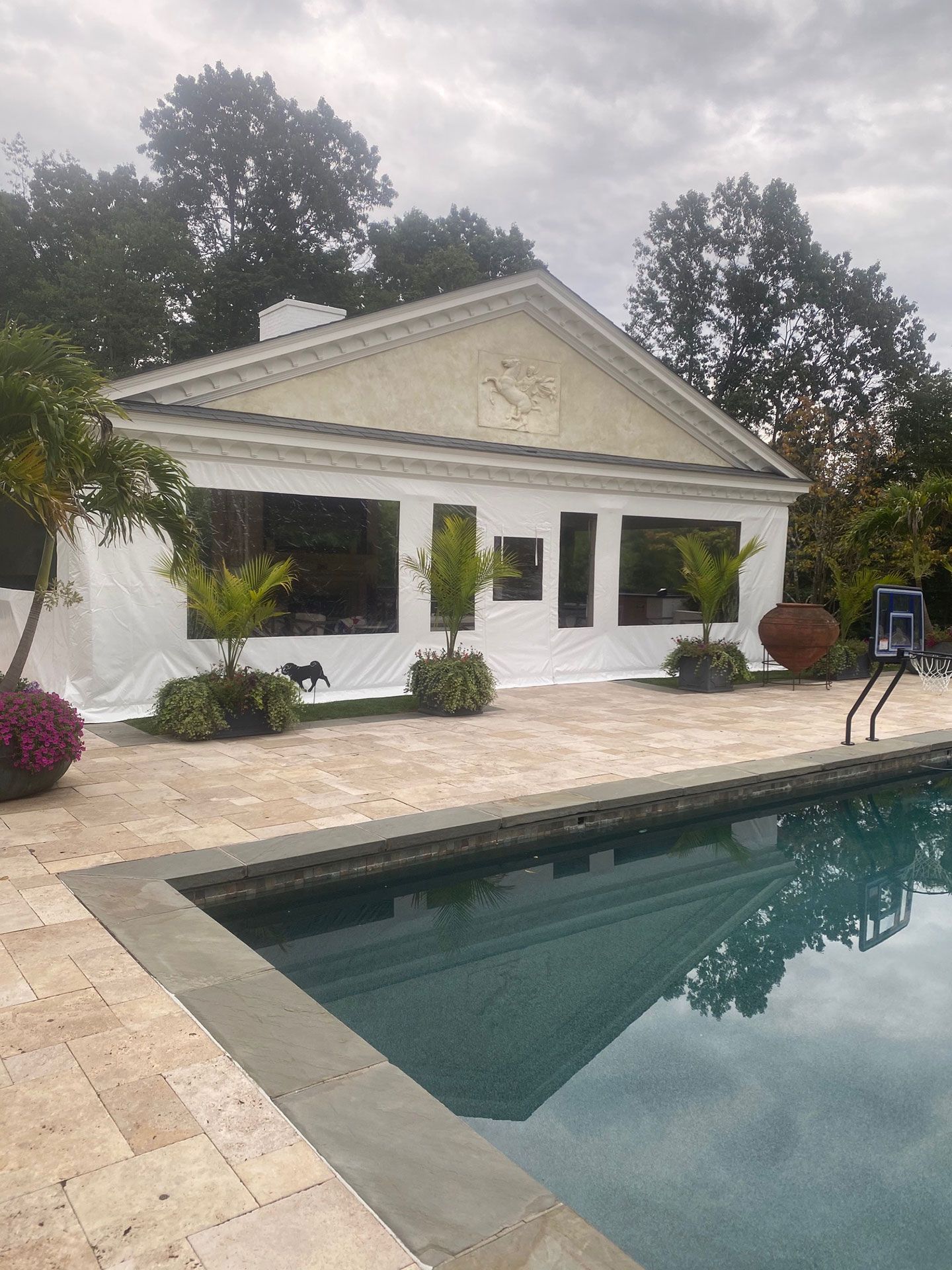 Poolside building with large windows, beige facade, and a pool in the foreground, surrounded by greenery, overcast sky.