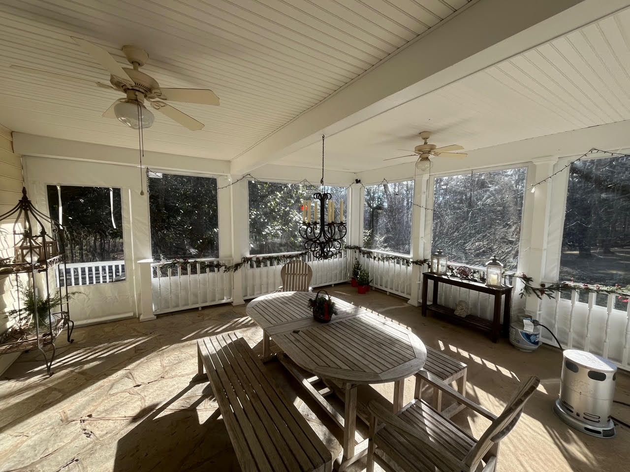 Enclosed porch with clear curtains, wooden furniture, ceiling fans, and a chandelier; sunlight streams in.