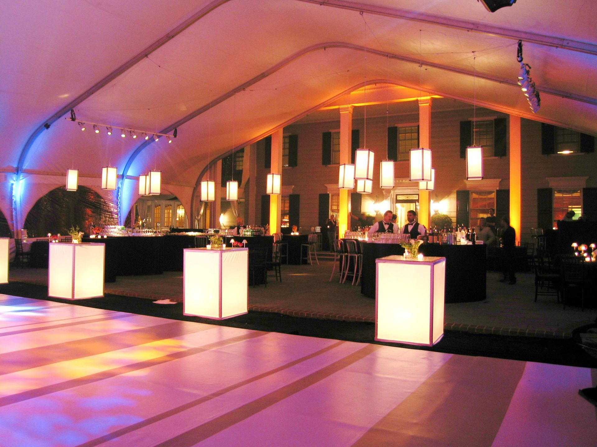 Event tent with lit dance floor and tables, outside a building at dusk, purple and orange lighting.