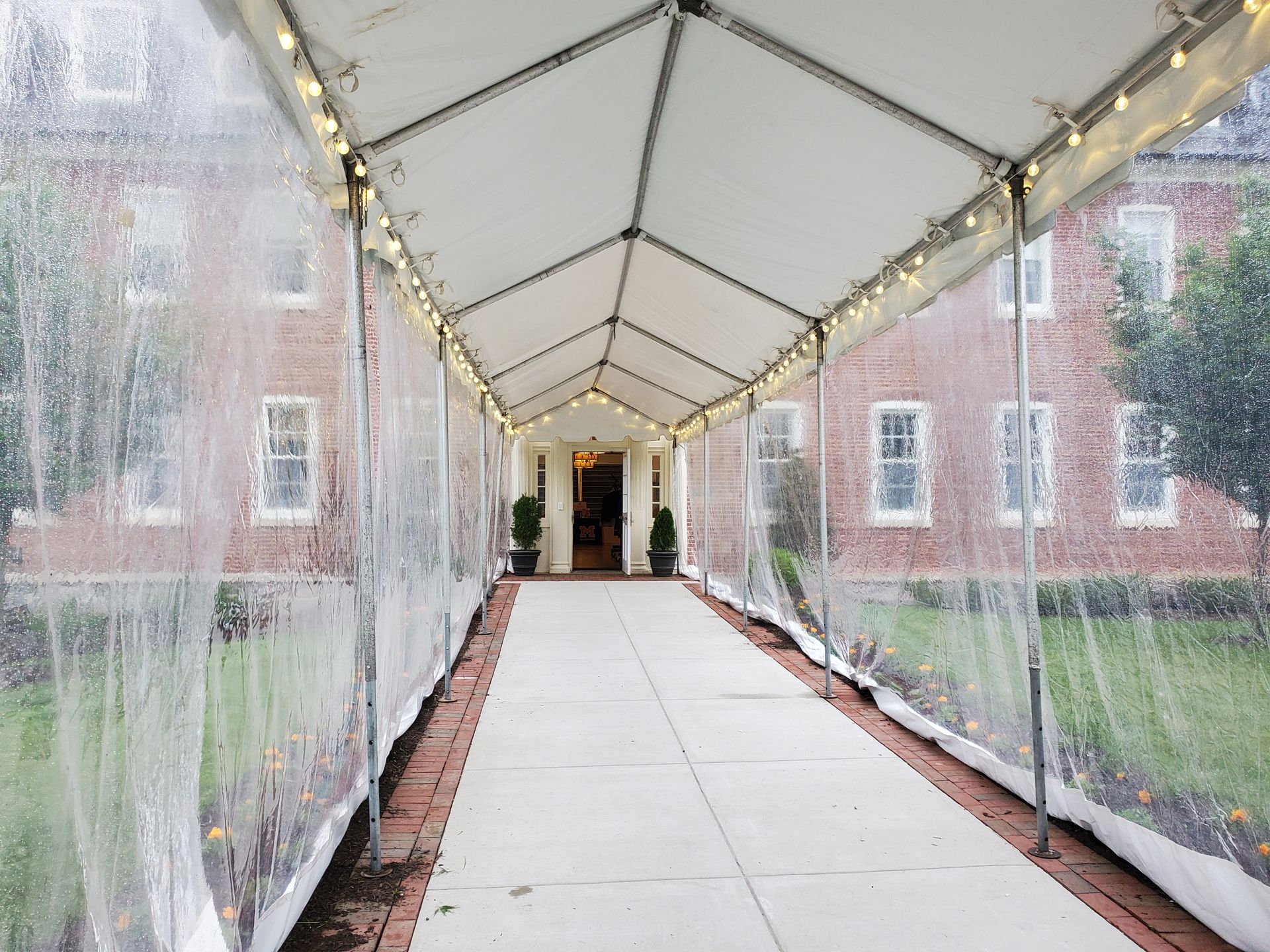 Covered walkway with clear sides leads to a brick building entrance, with string lights overhead.