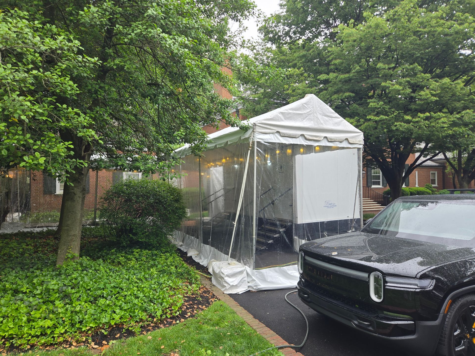 A white tent with clear plastic walls attached to a house, next to a black SUV.