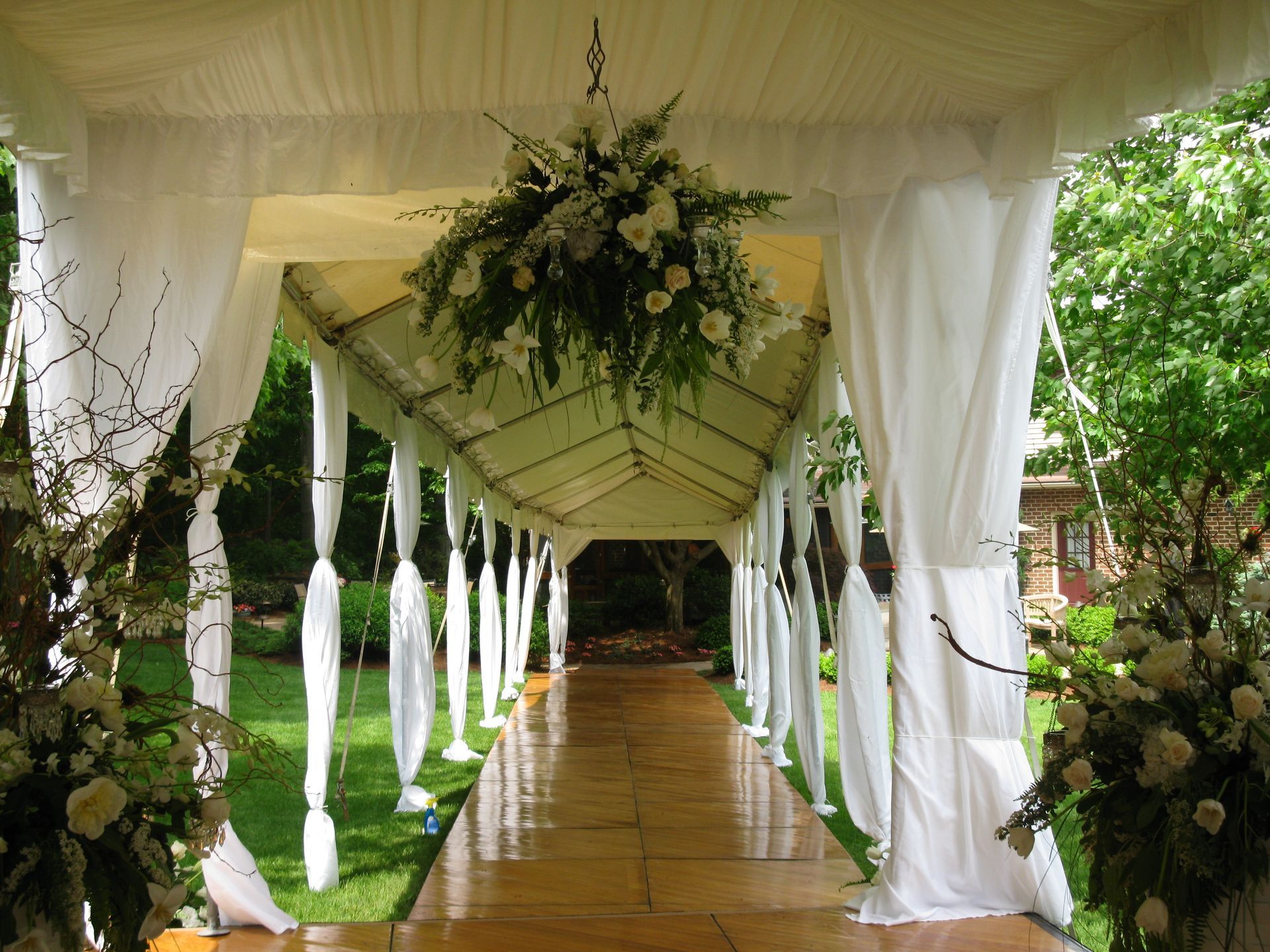 Covered walkway decorated for an outdoor event with white draping, floral arrangements, and a wood floor.