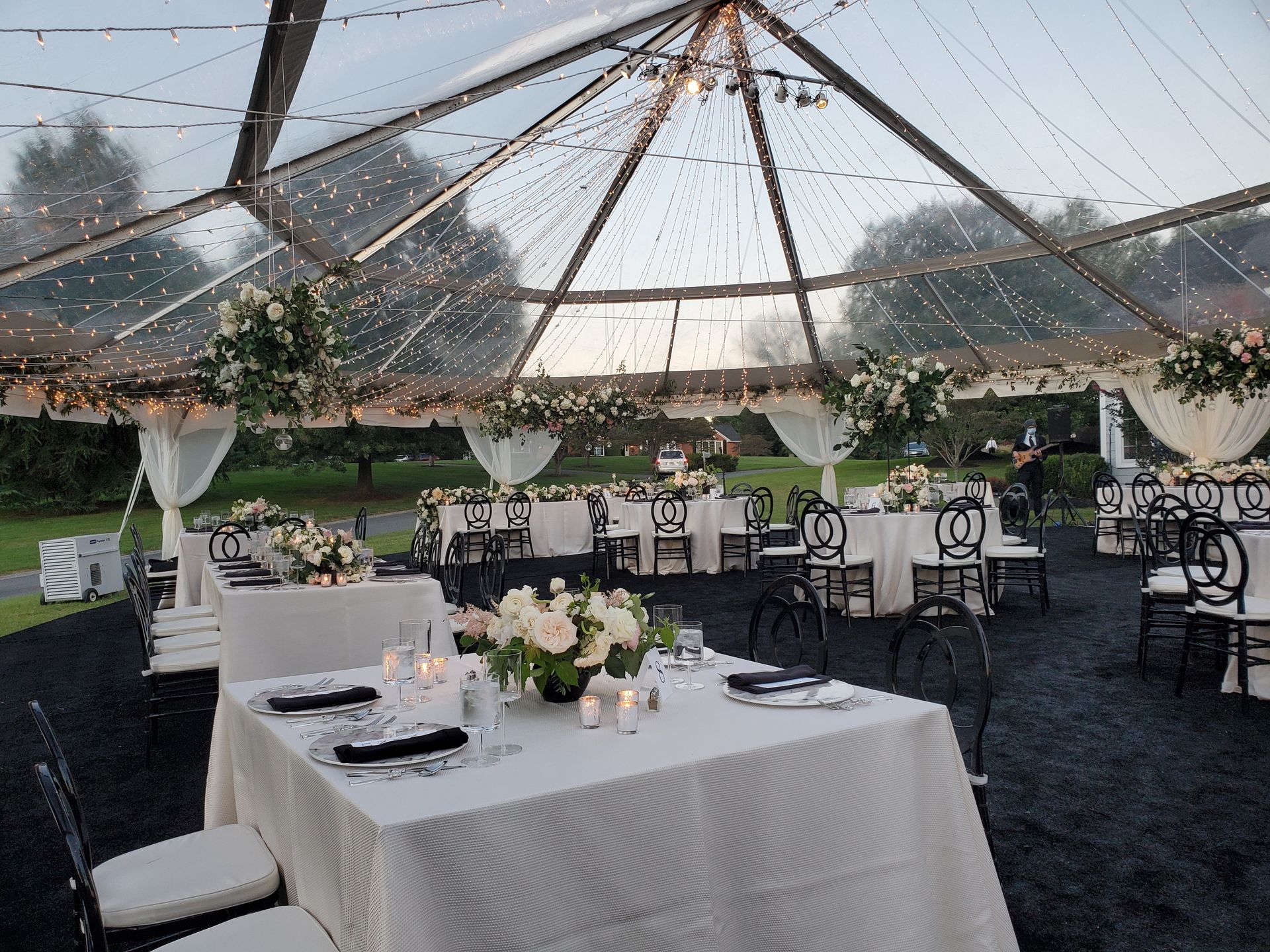 Wedding reception setup inside a clear tent; tables set with floral arrangements, black chairs, and string lights.