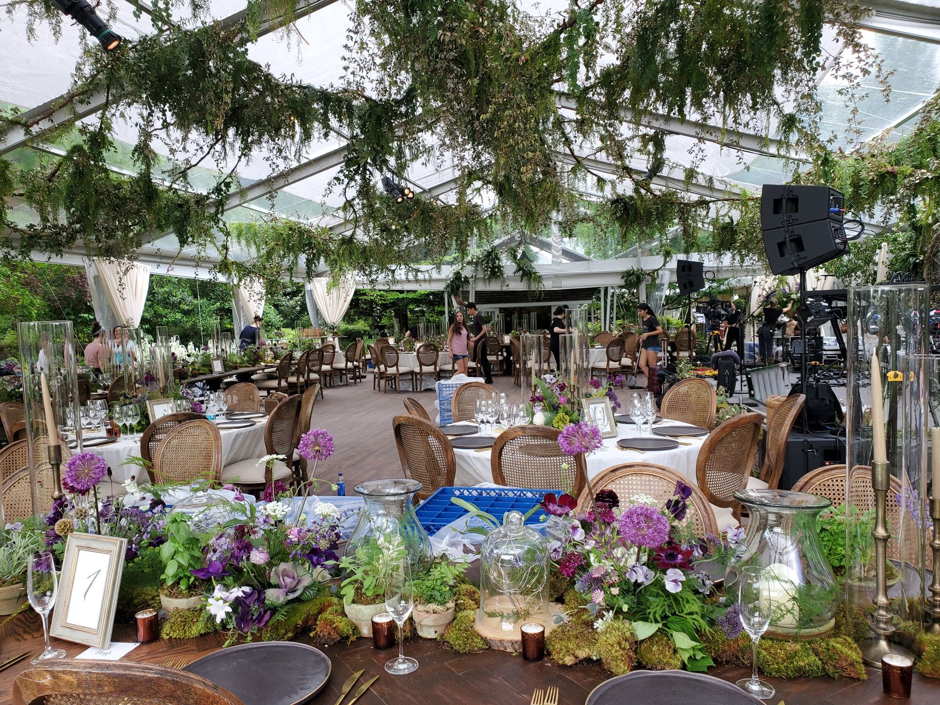Decorated event space, tables set with flowers and linens under greenery-covered tent.