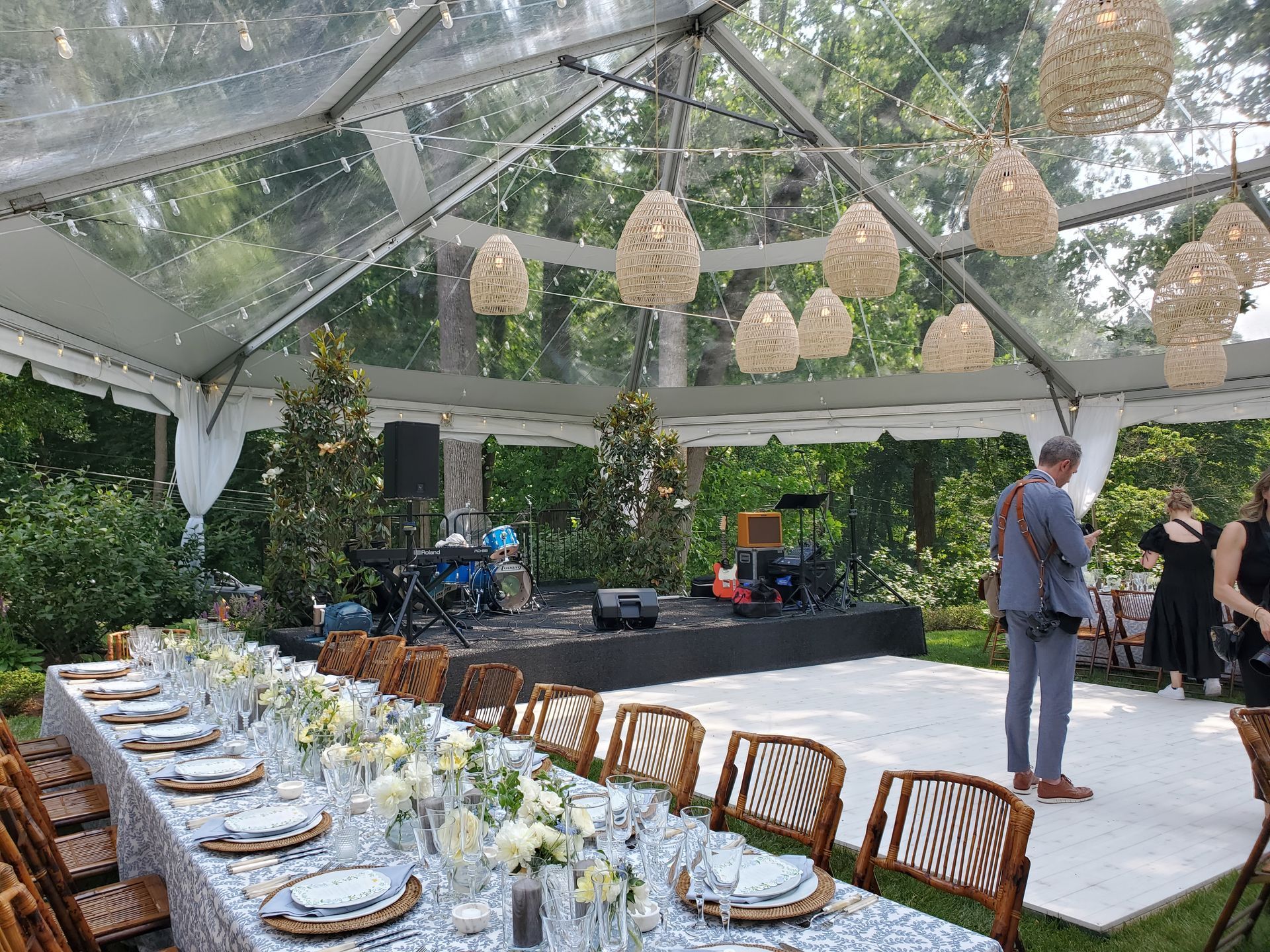 Outdoor event with clear tent, long table set for dinner, stage with band, and hanging lights.