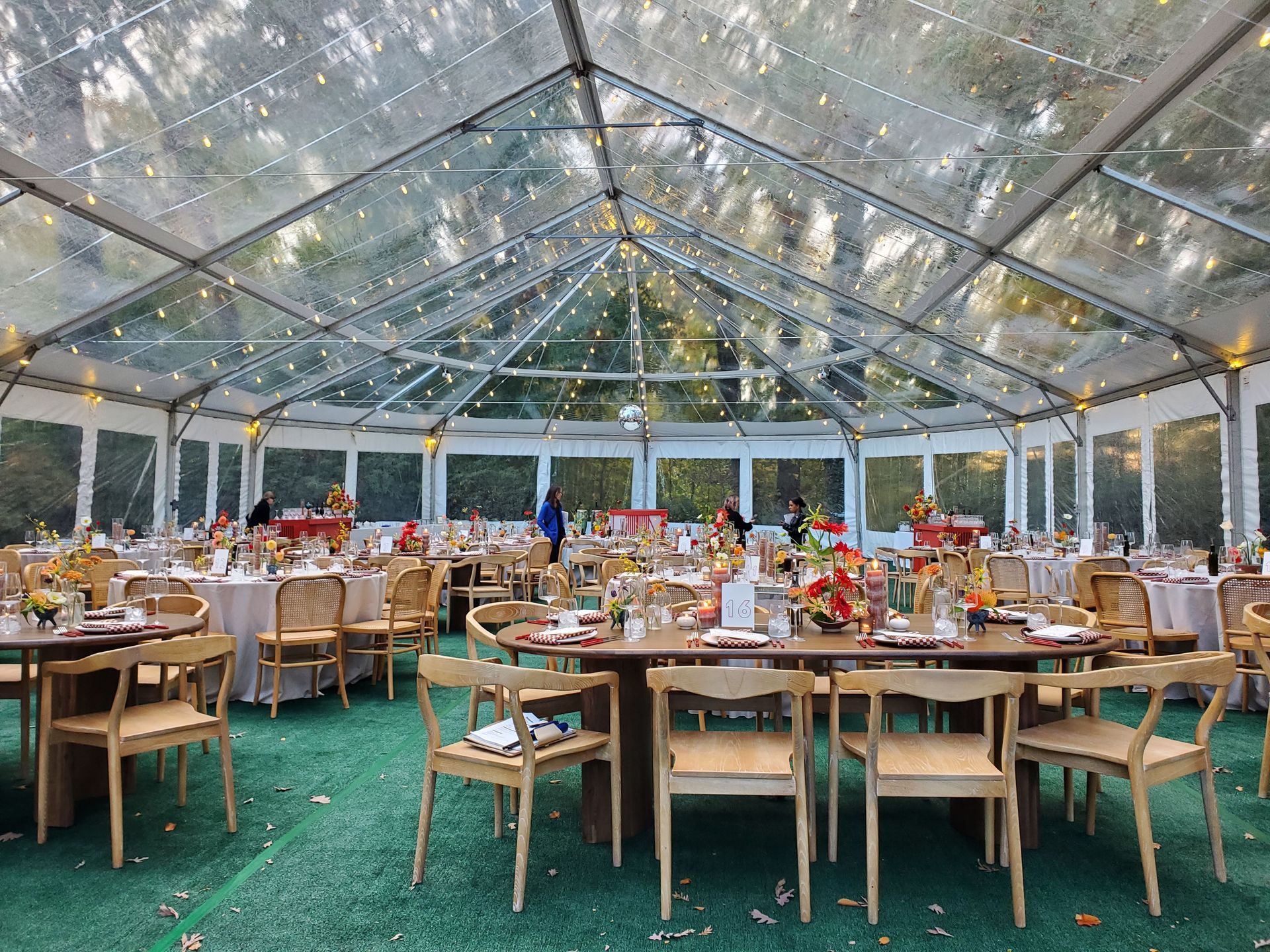 Clear tented event space with round tables, decorated for a gathering, with string lights and trees visible.