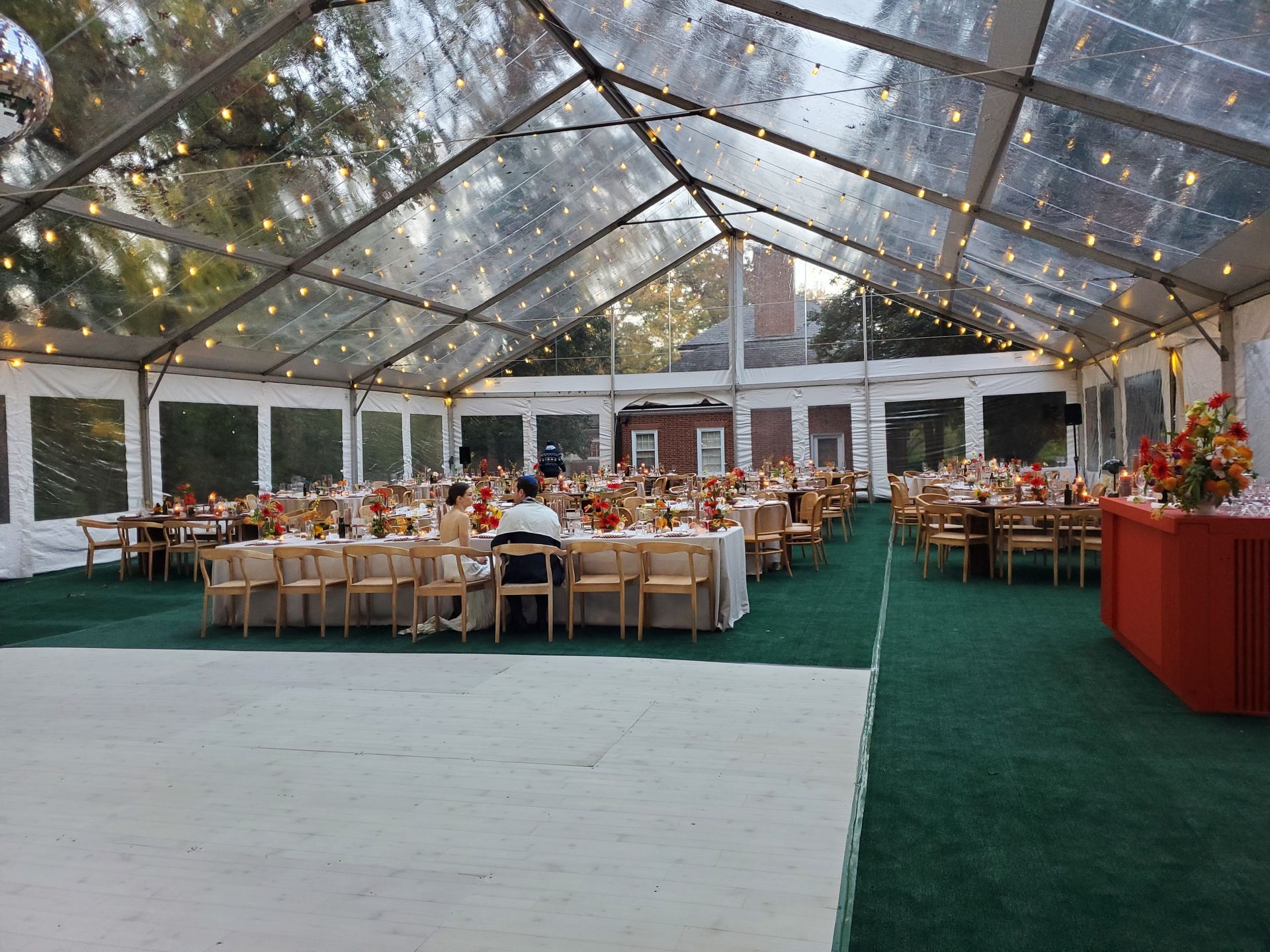 Clear tented event space with tables set for a gathering, a dance floor, and string lights.