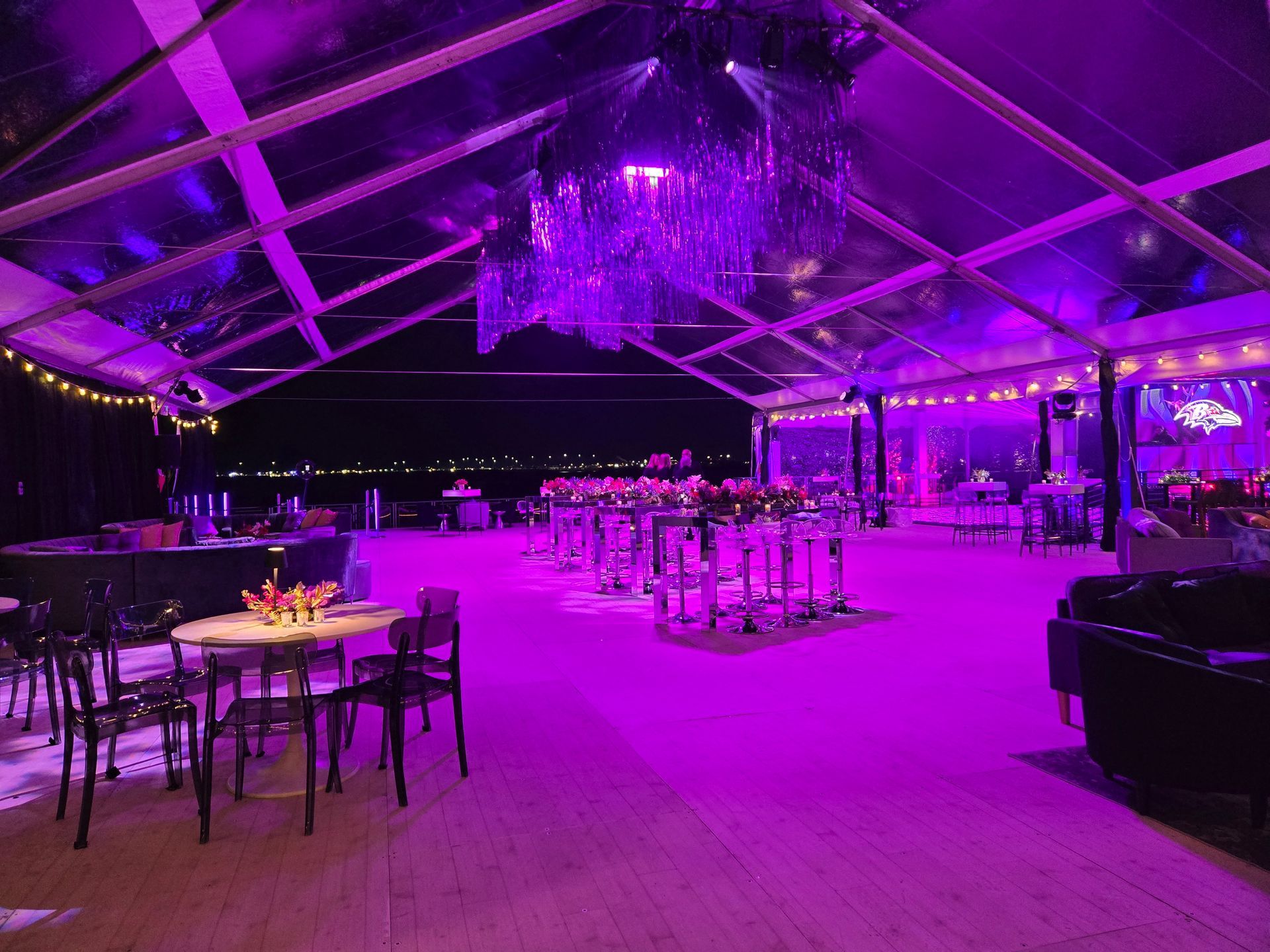 Purple-lit event tent interior with tables, chairs, and bar, overlooking a dark body of water.