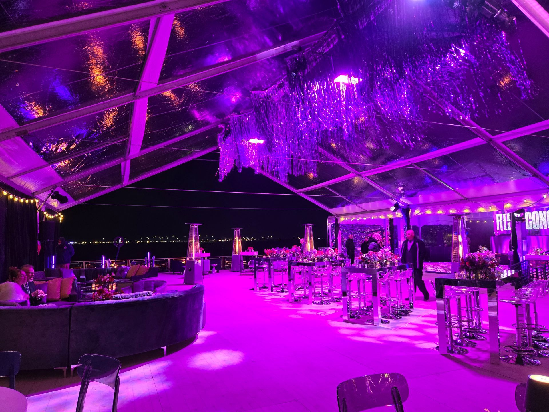 A clear tent lit in purple, with tables, people, and a bar overlooking a dark body of water.
