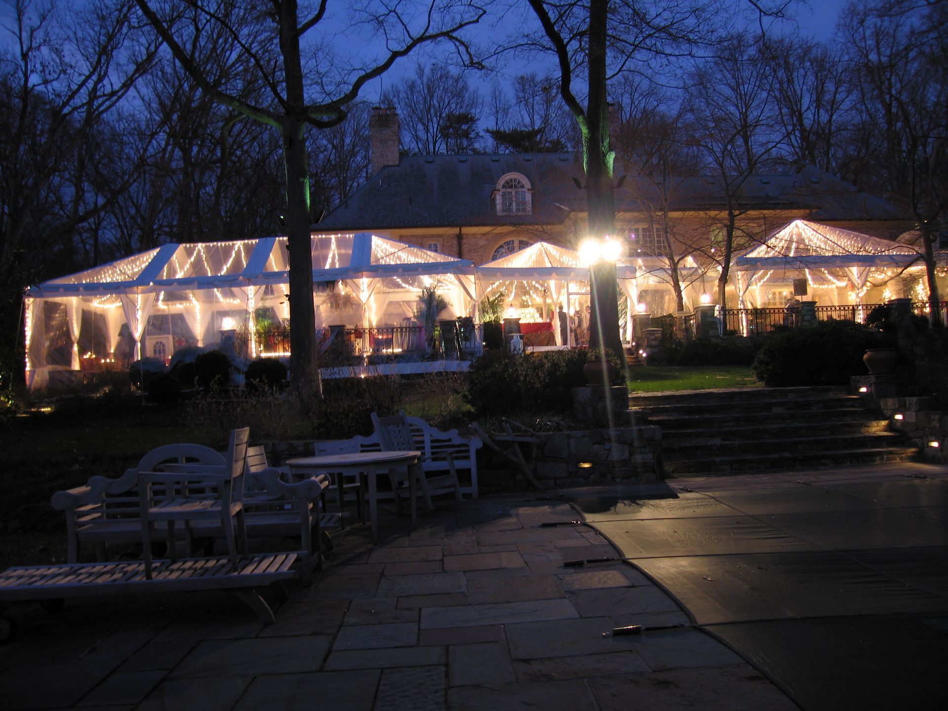 Outdoor event lit up at night with a transparent tent and dining tables. Building in the background.