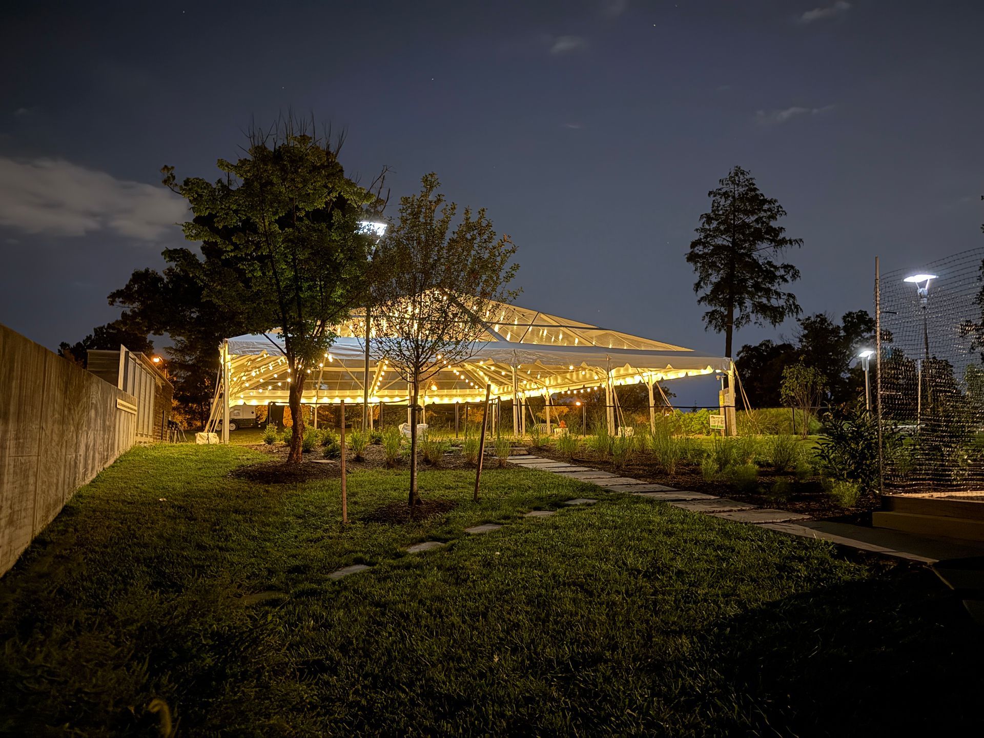A nighttime scene with a gazebo lit by string lights in a garden, grass in foreground, trees and a wall.