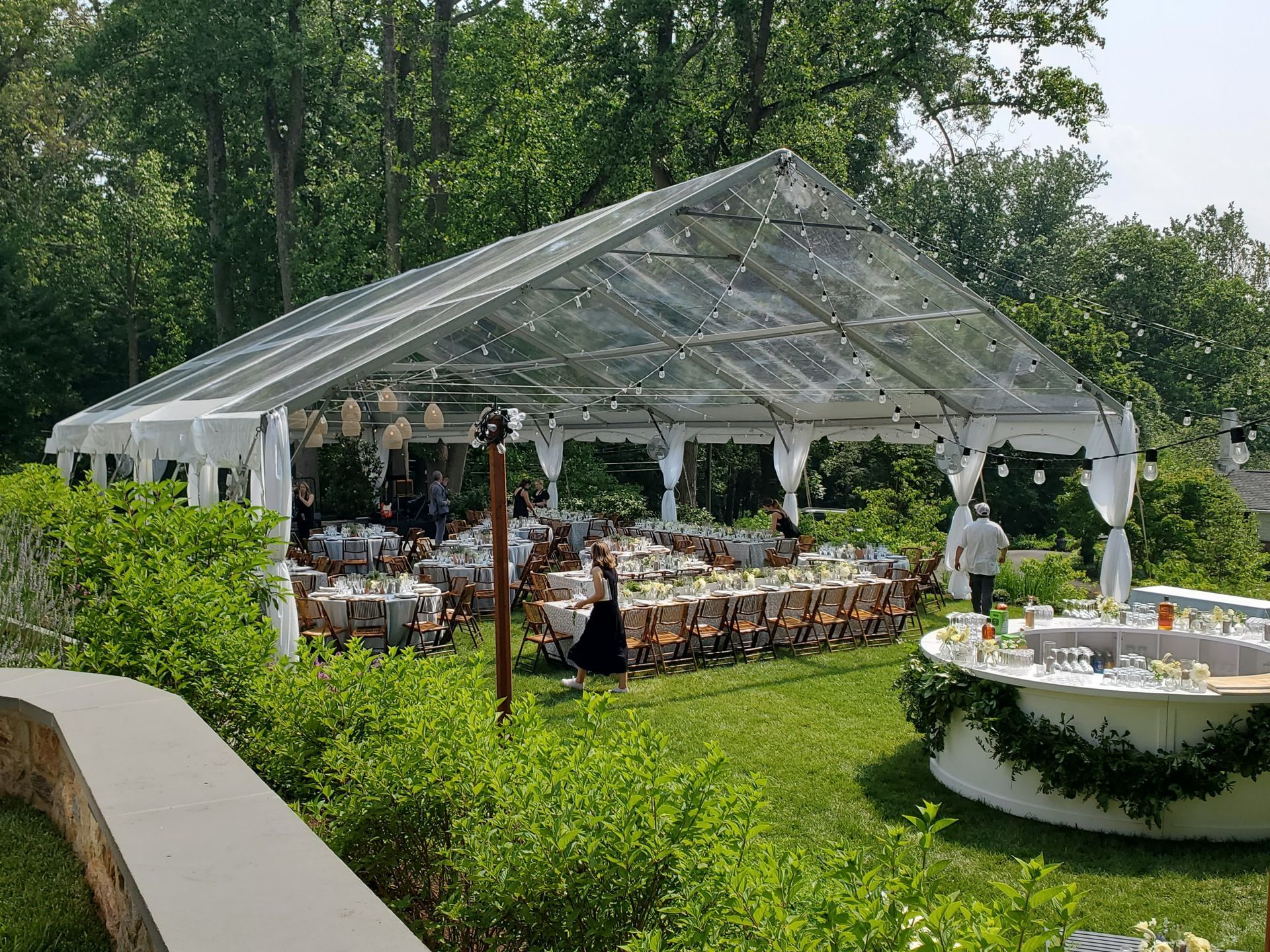 Clear tent set for an outdoor event; tables set for guests; person walking by; surrounded by greenery.