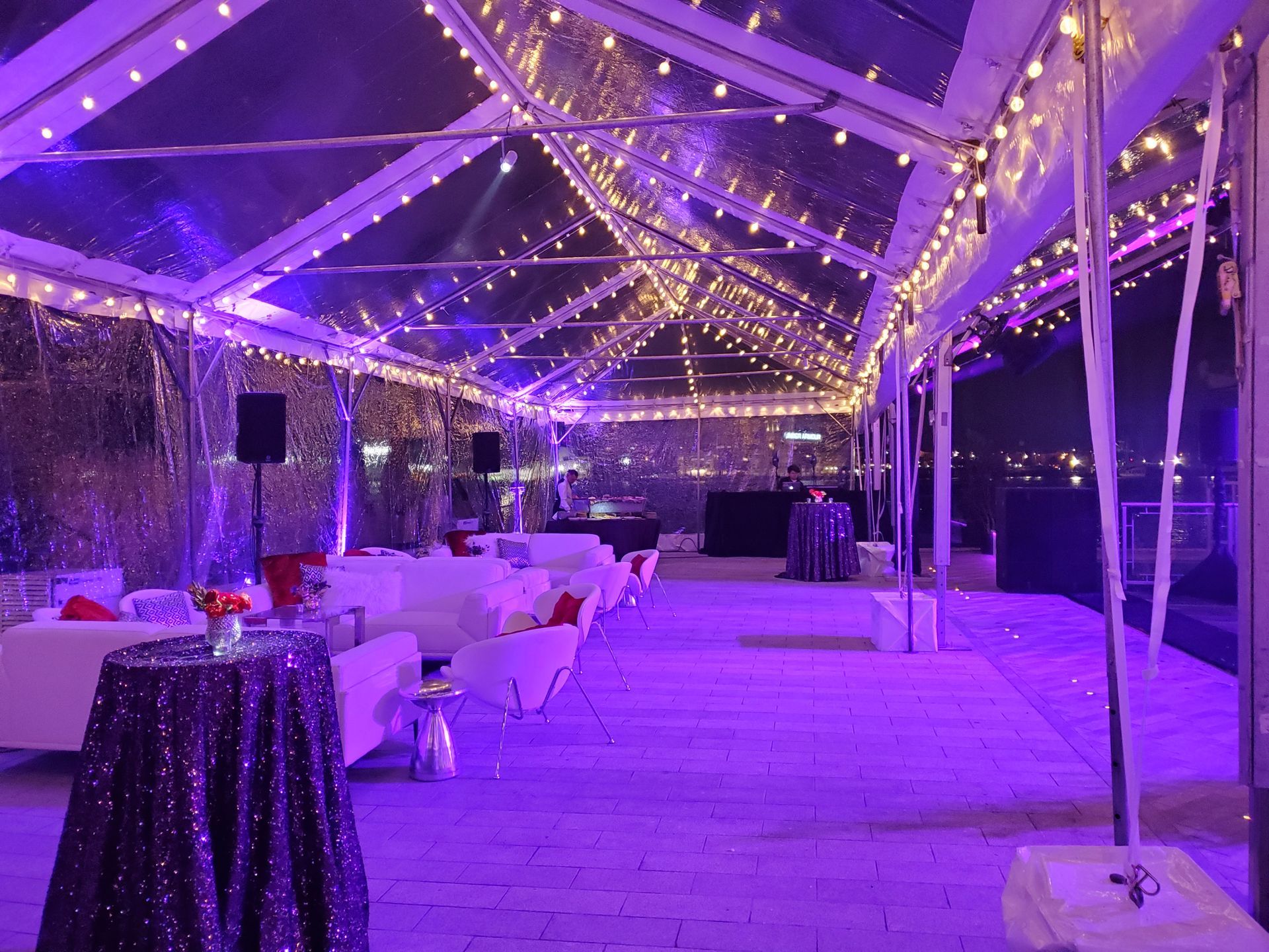 Clear tent lit with purple lights, small tables, white couches, and a city view at night.