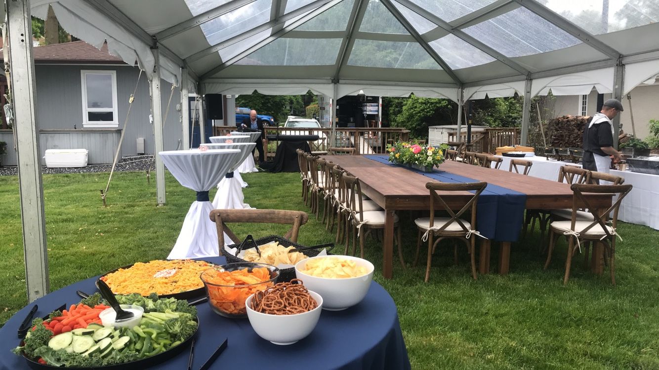 Outdoor event with food tables, dining tables, and a clear tent in a grassy backyard.