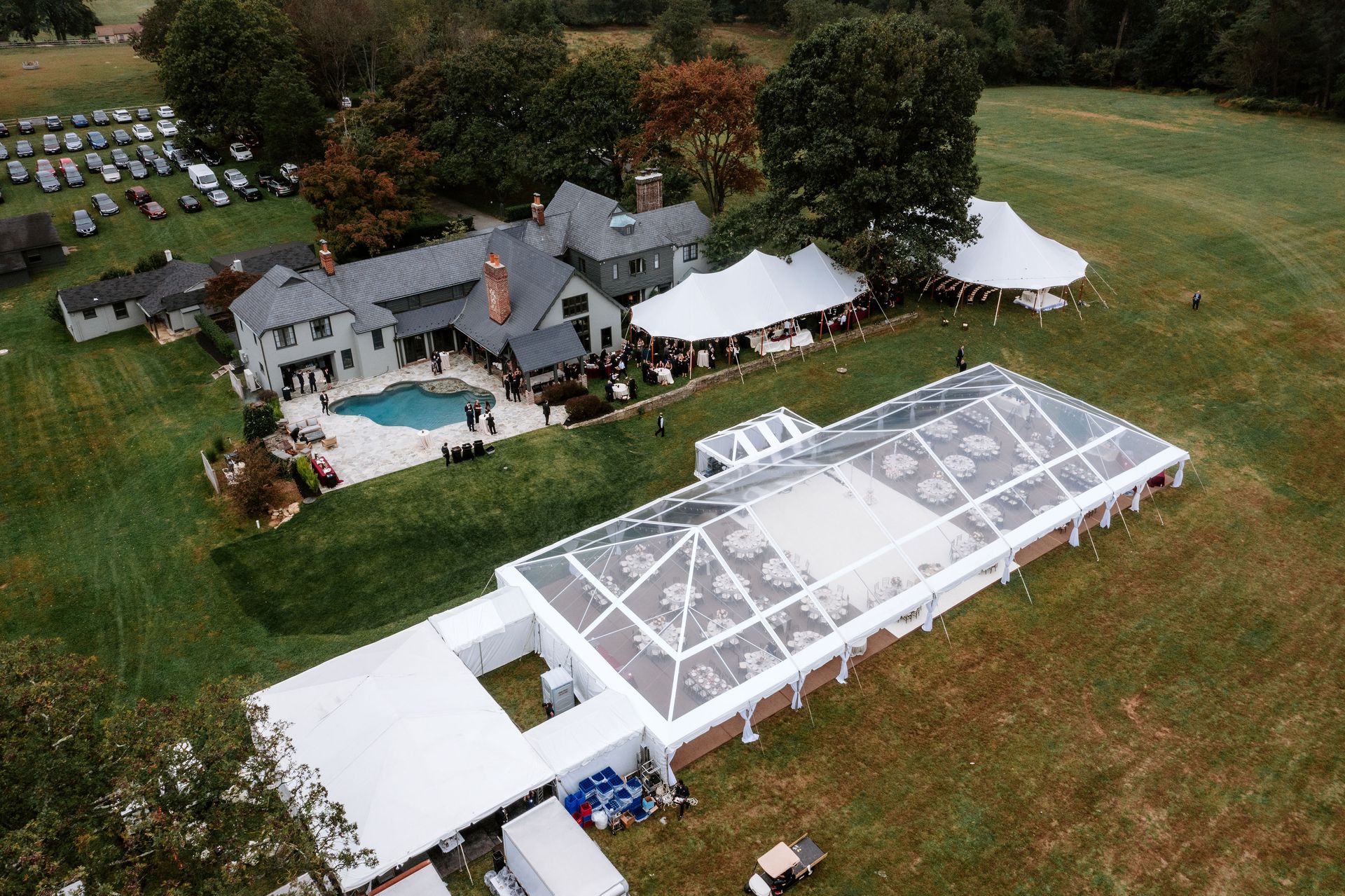 Aerial view of a house with a pool, multiple tents on a grassy field; an event taking place.