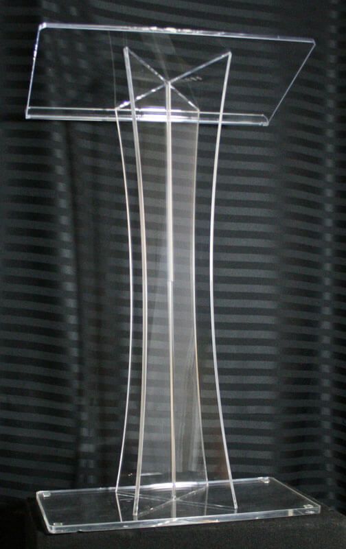 Clear acrylic podium on a black backdrop, featuring a sloped top and base with a flared central support.