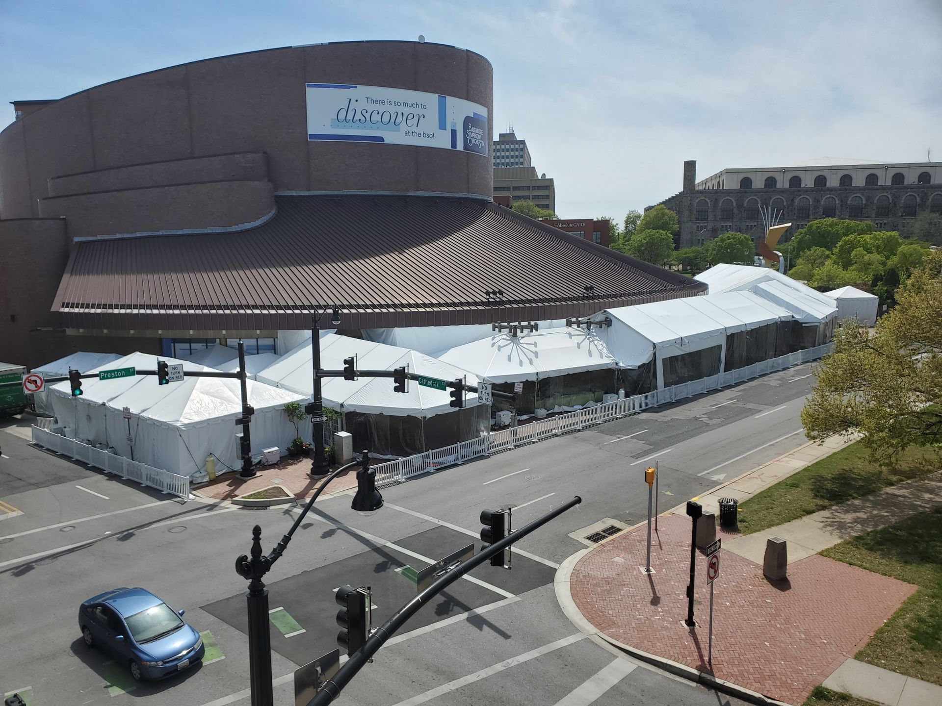 Building with brown roof and white tents set up along a street corner in front of it. Blue car on street.