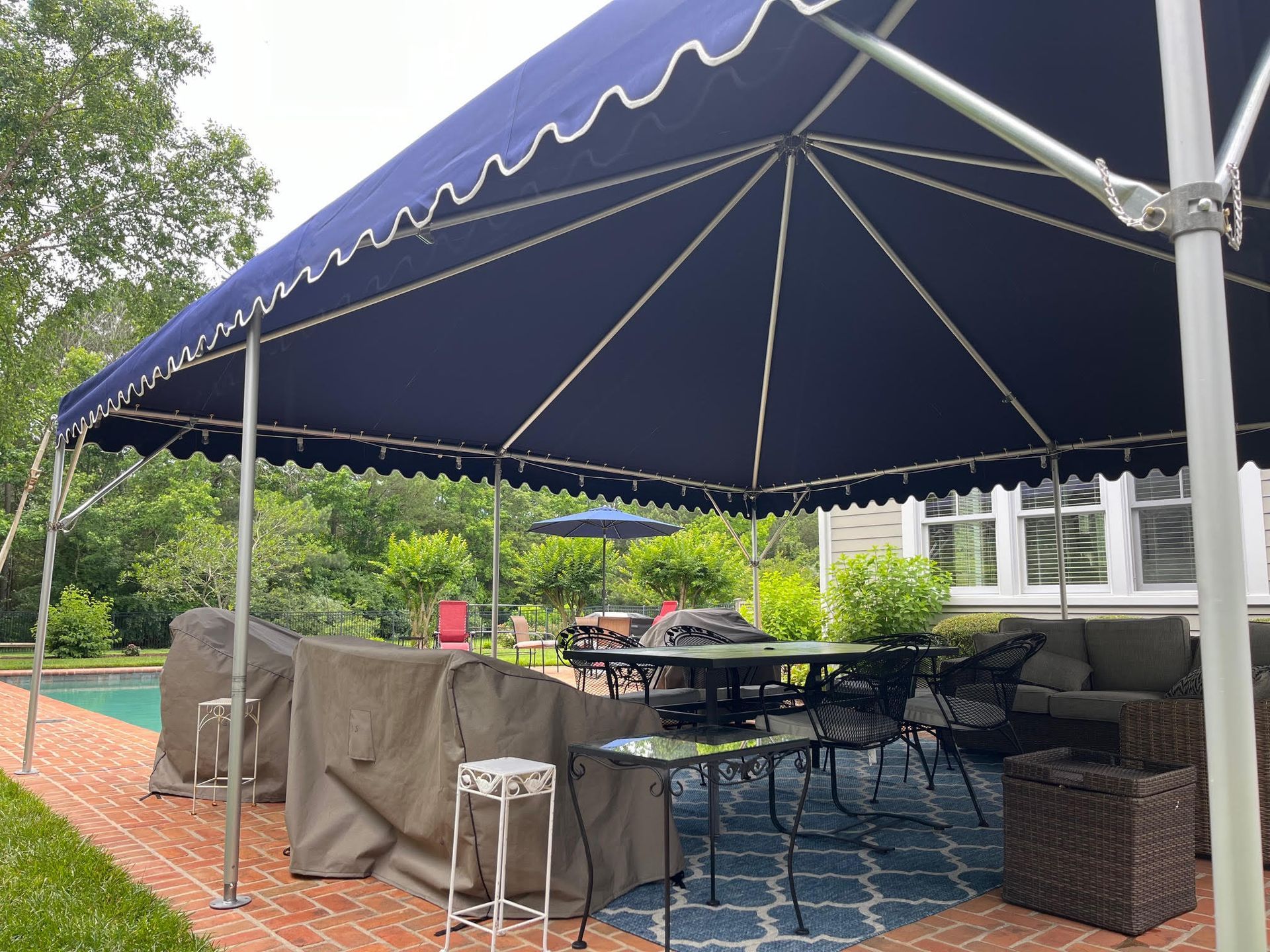 Patio with navy blue canopy over a dining table and furniture. Brick patio, green trees, and a pool in the background.