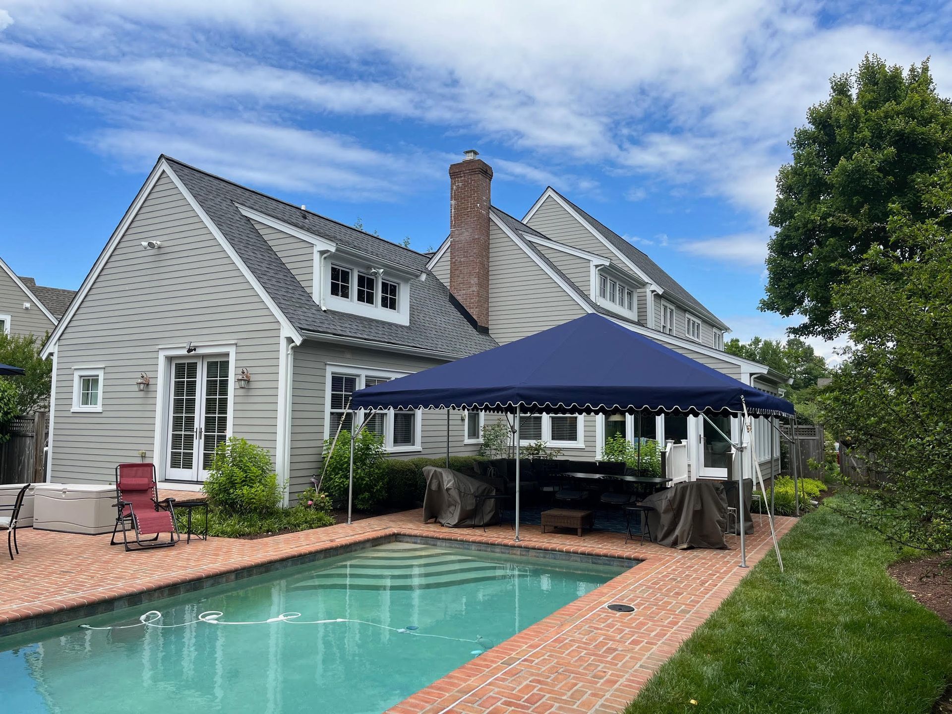 Backyard pool with canopy, house, brick patio, and green lawn under a blue sky.