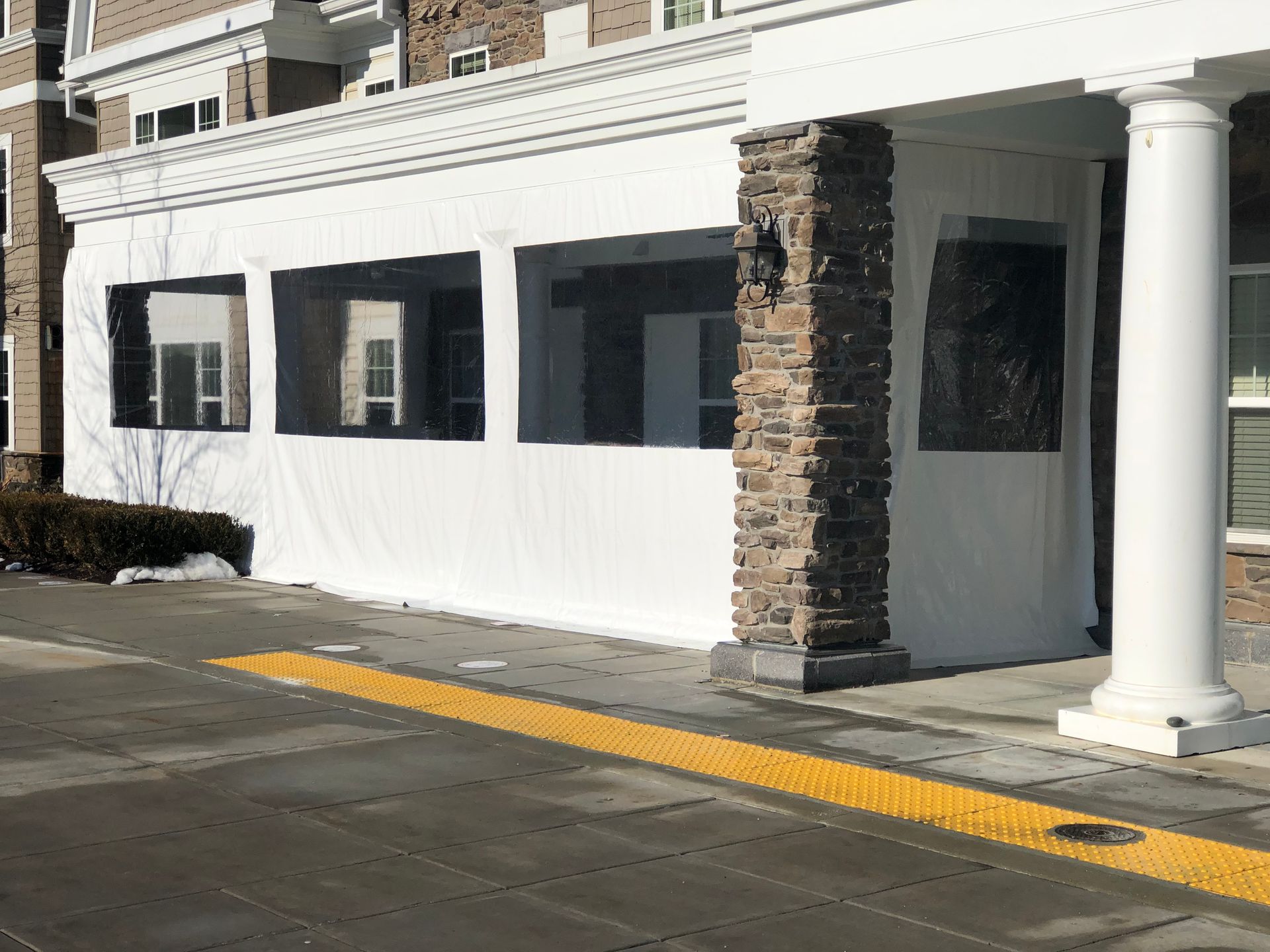 White enclosed outdoor seating area with clear plastic windows; building with brick and white columns.