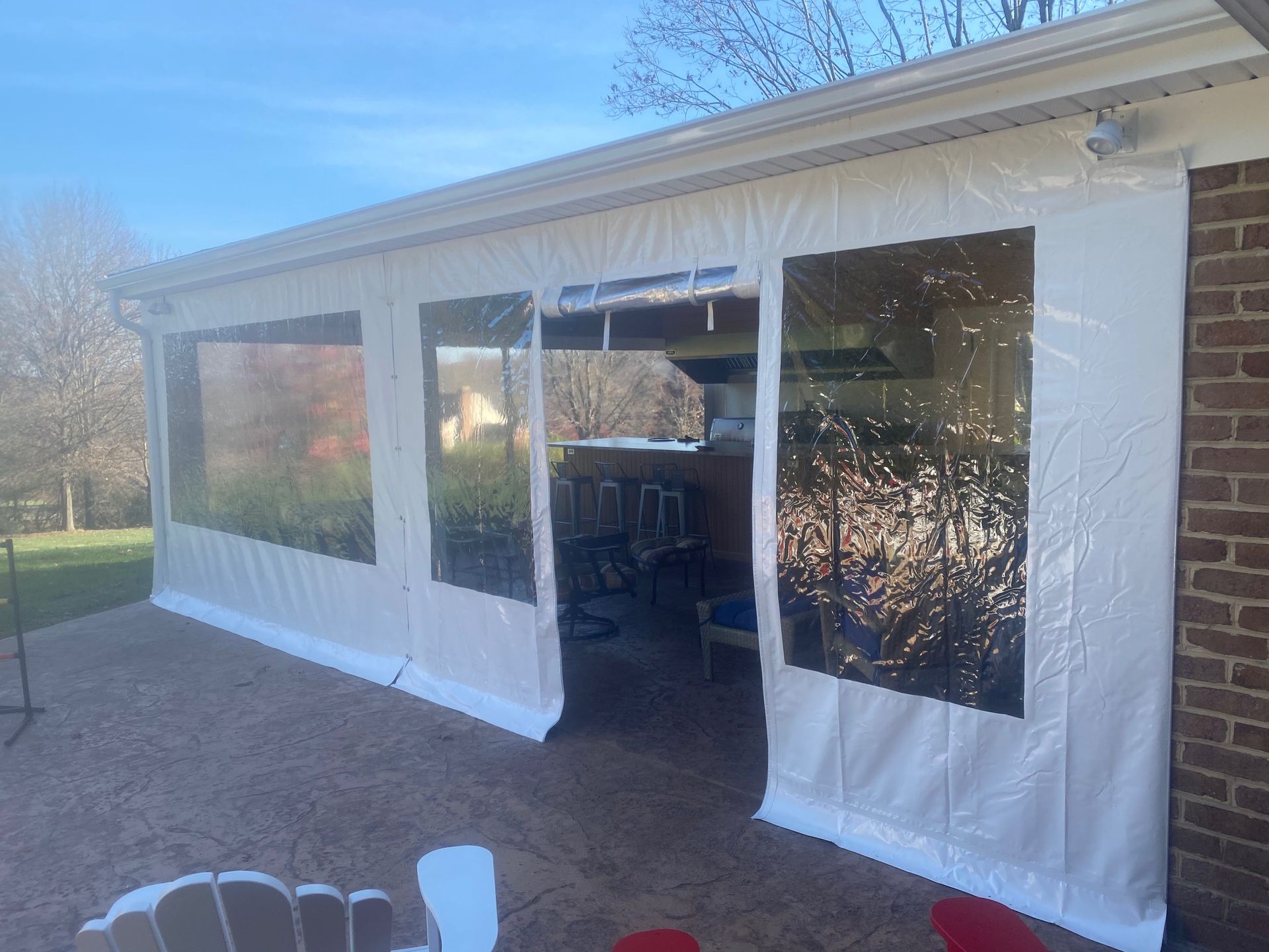 Outdoor patio with white curtains and clear vinyl windows attached to a brick house, sunny day.
