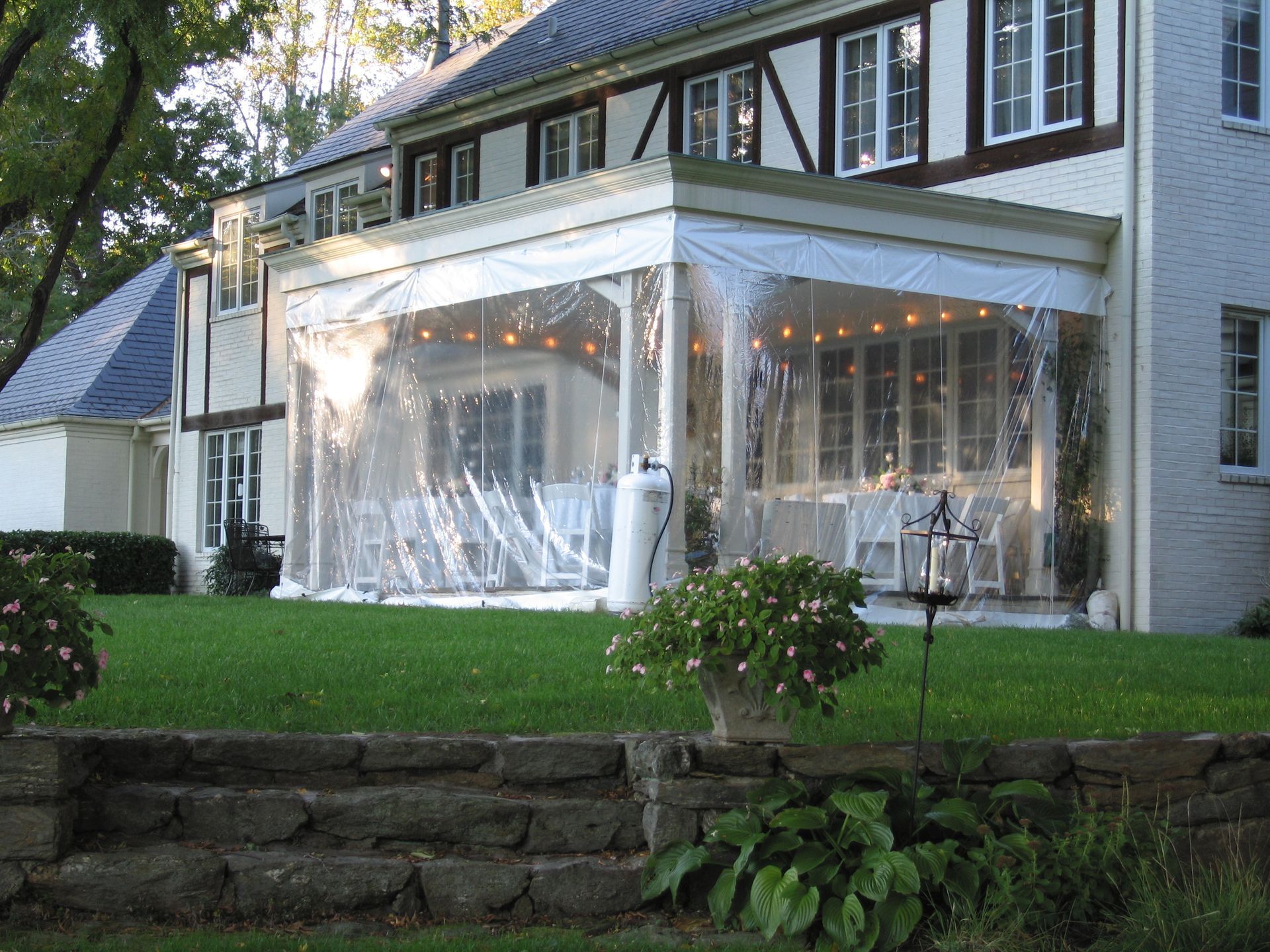 White house with clear plastic enclosed porch, string lights, and a lawn.