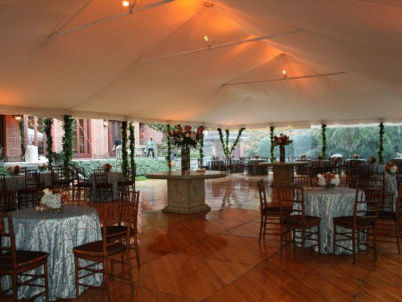 Inside a large white tent, tables with silver tablecloths and wooden chairs are set for an event.