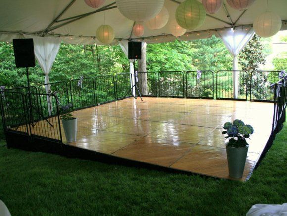 Wooden dance floor with black railing and speakers, under a tent, surrounded by grass and trees.