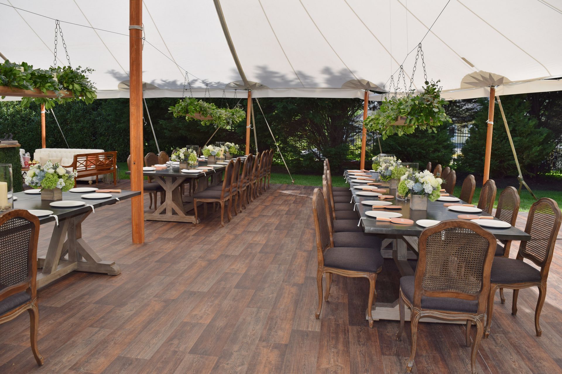 Outdoor dining tables under a white tent, set for a gathering. Wooden tables with chairs, floral centerpieces.