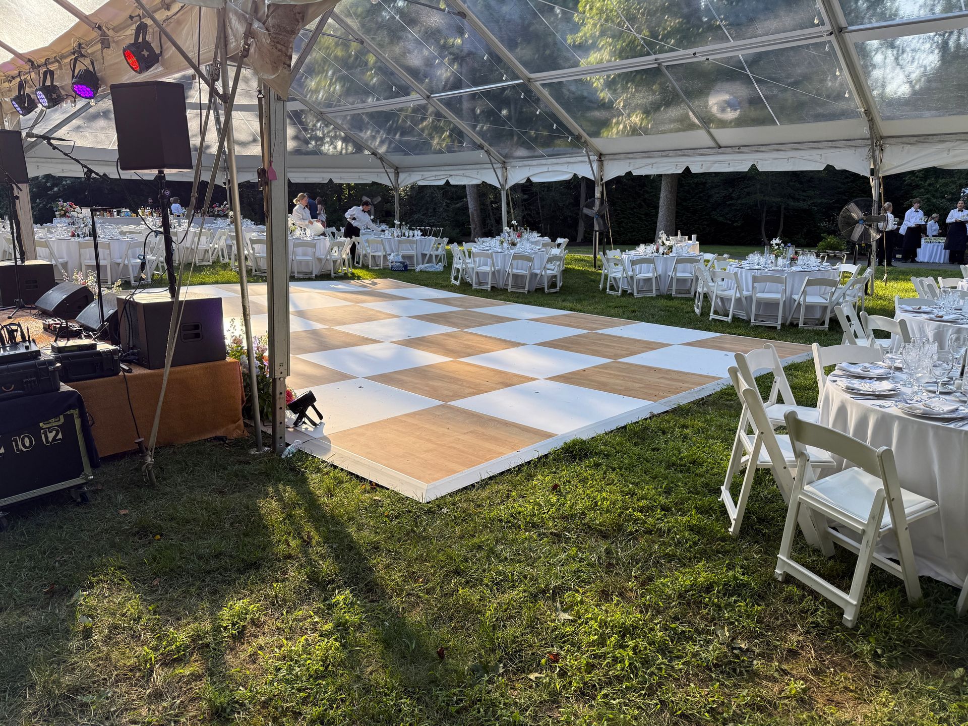 A wedding reception setup under a clear tent with tables, chairs, a dance floor, and speakers in a grassy area.