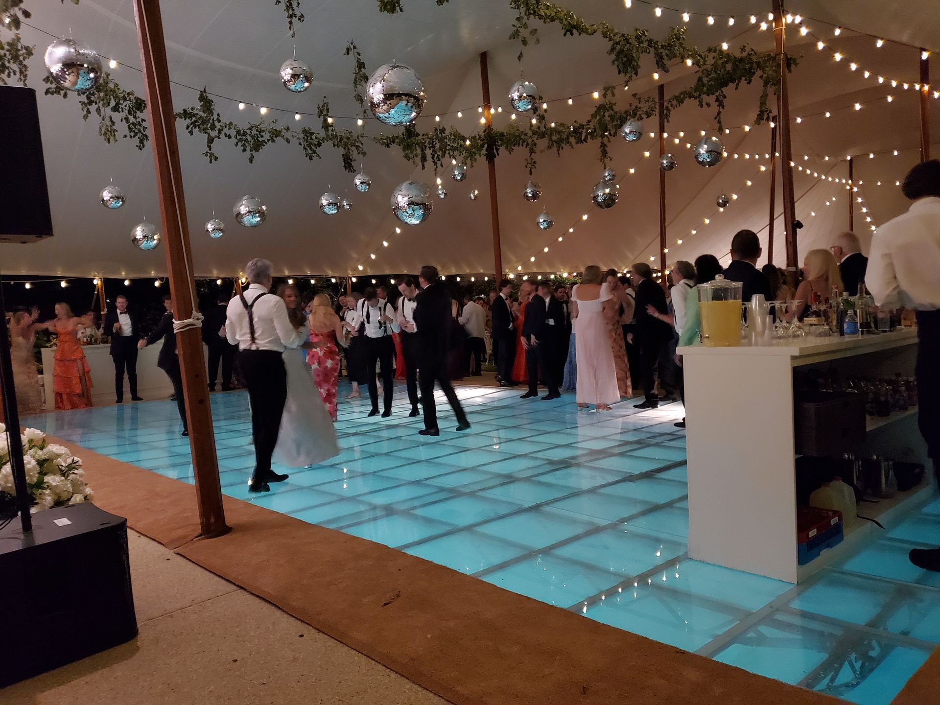 People dancing on a lit, glass dance floor in a tent decorated with lights and disco balls.