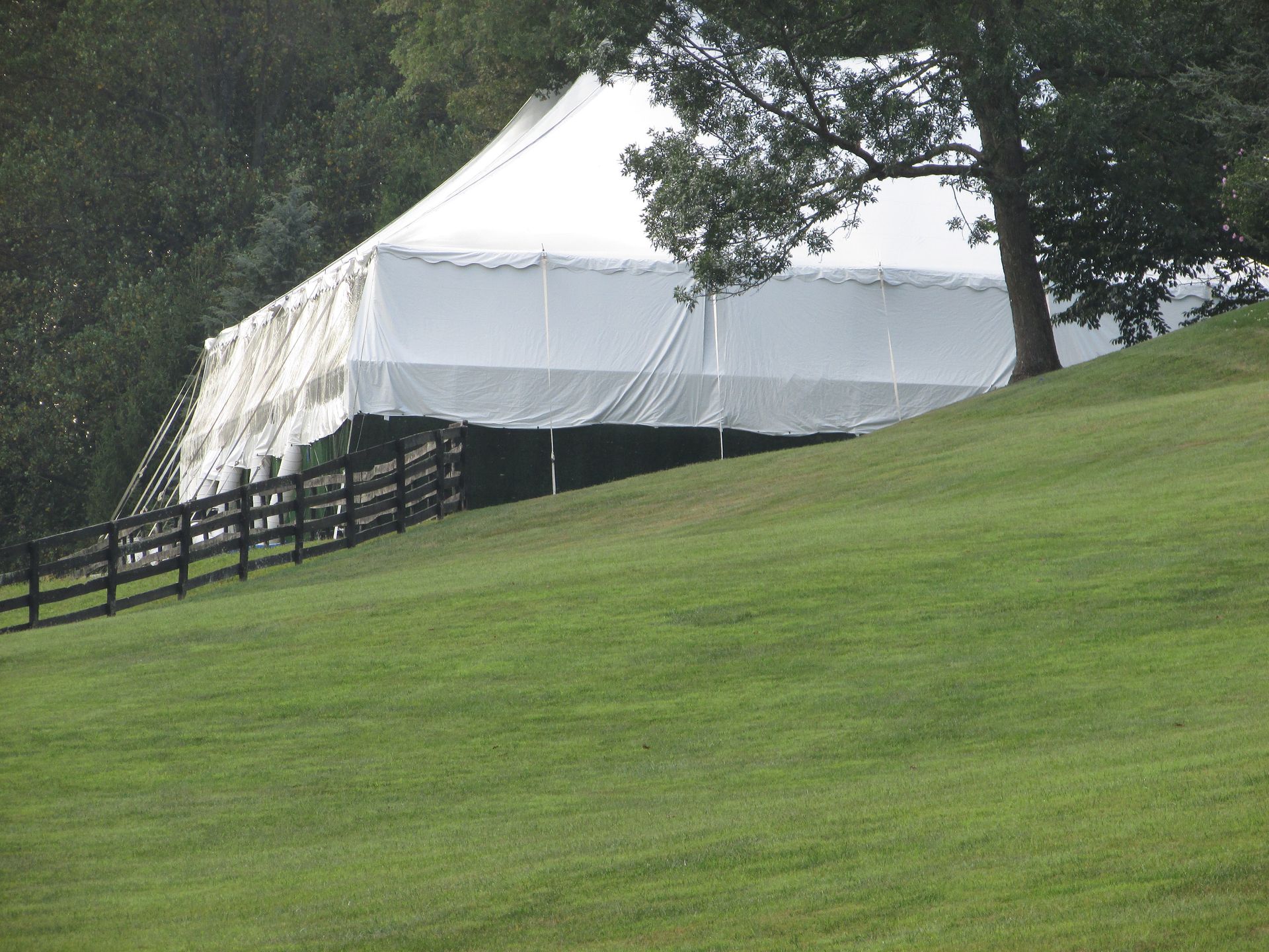 White tent on a grassy hill next to a tree and a wooden fence, under a rainy sky.