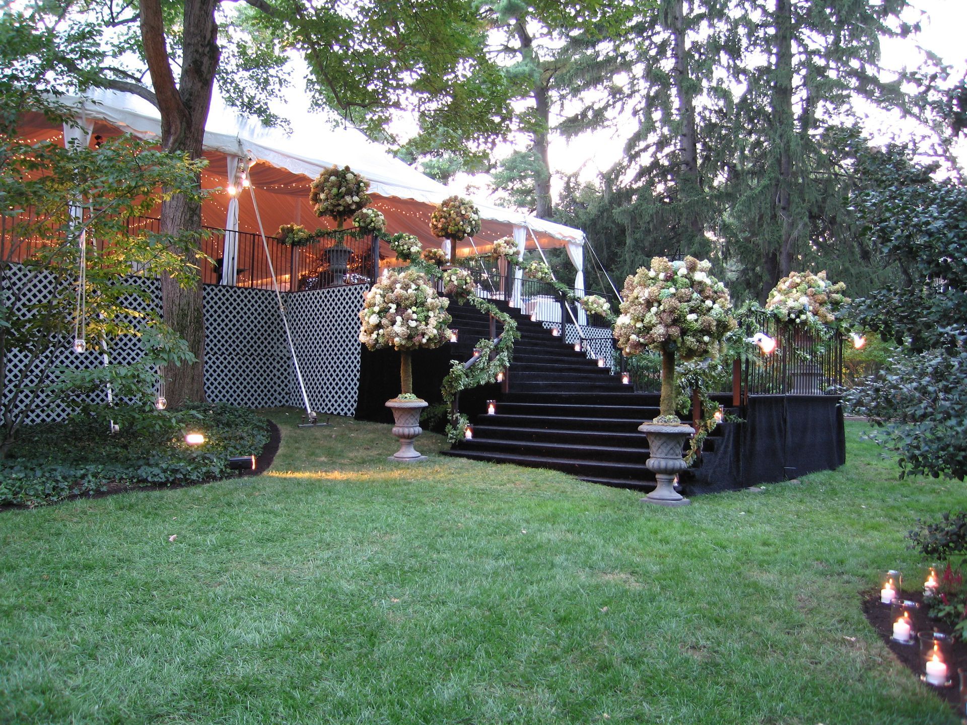 Black staircase leads to white tent, flanked by floral arrangements, in a garden setting.