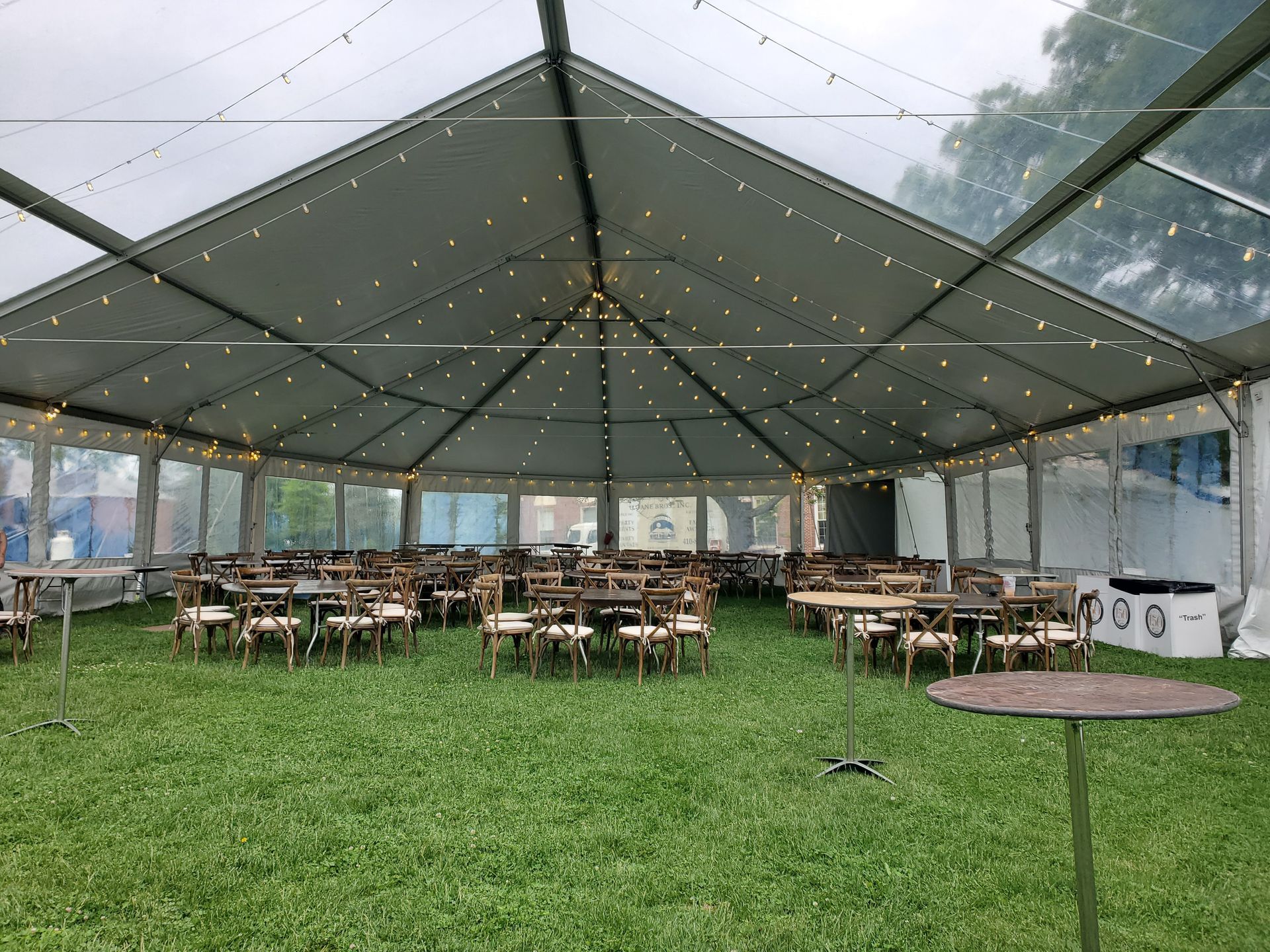 Inside a clear tent: tables and chairs on green grass, strung lights.