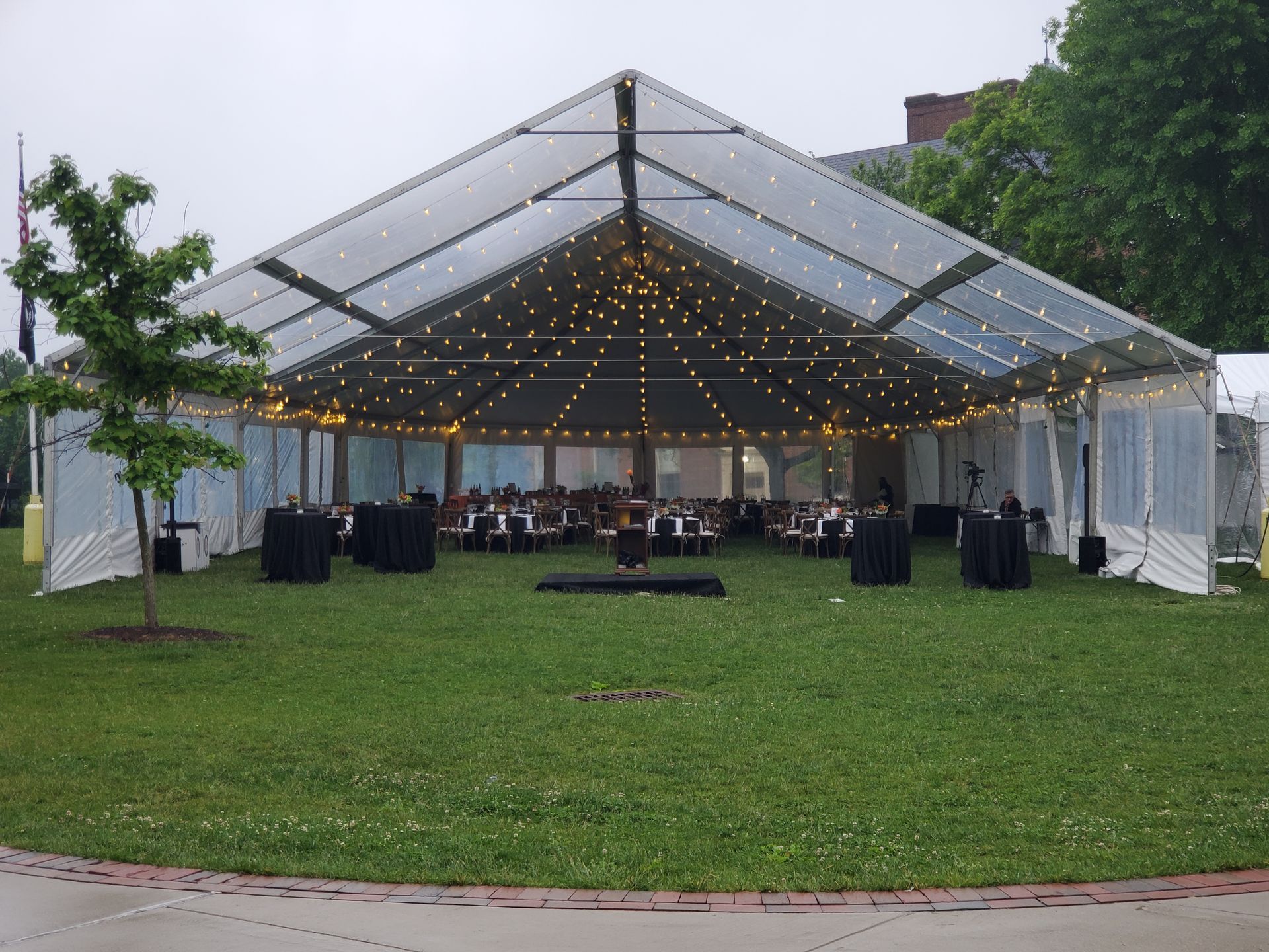 Clear tent set up on green grass for an event, with string lights, tables, and chairs.
